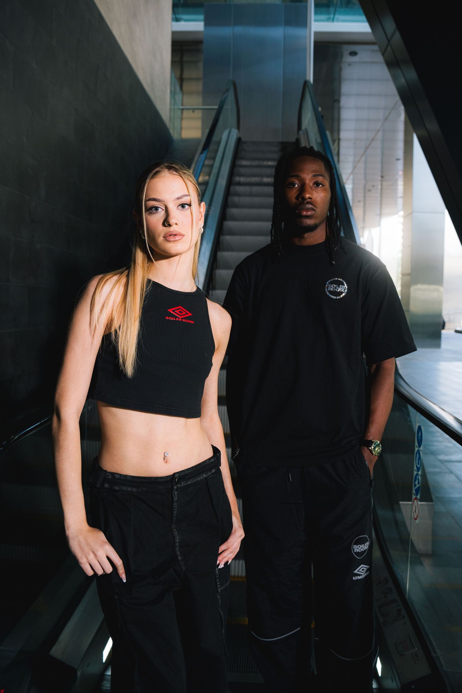 Athletic duo in black outfits pose against modern architectural backdrop with dramatic lighting.
