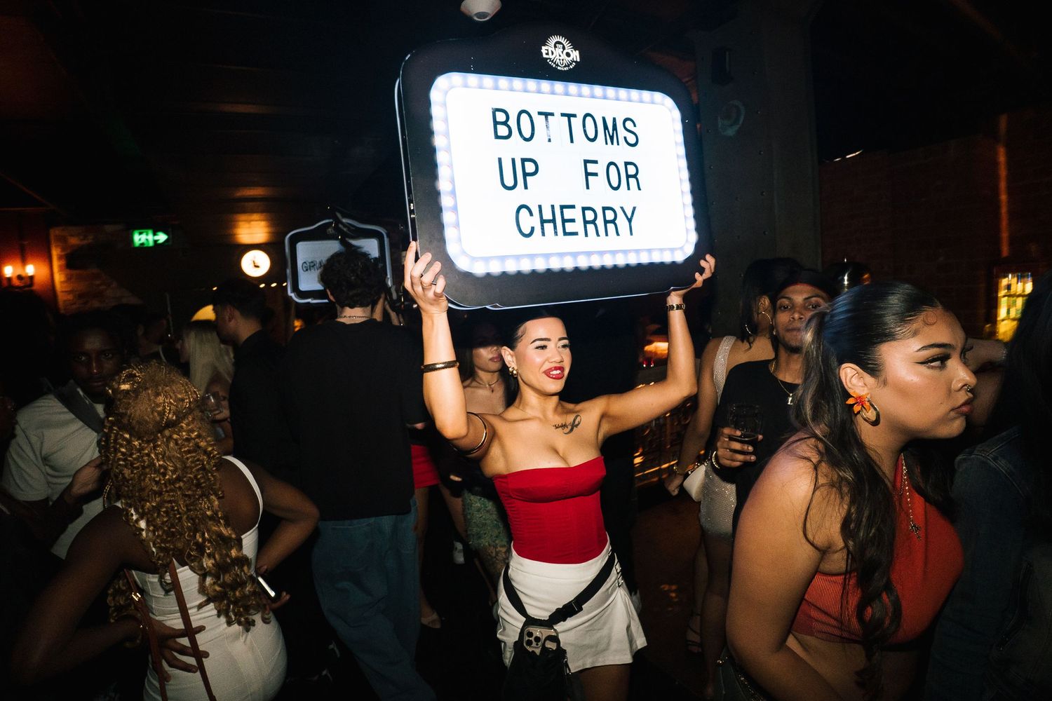 A crowd of partygoers at a nightclub with someone holding up a sign that reads 'Bottoms up for Cherry'.