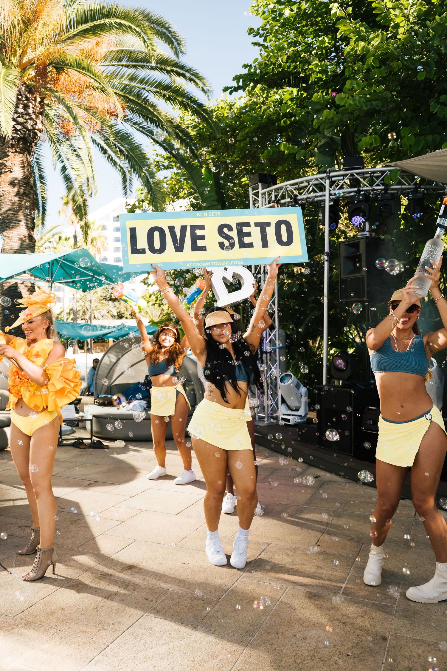 Dancers in colorful swimwear holding a 'LOVE SETO' sign perform at a tropical outdoor venue with palm trees.