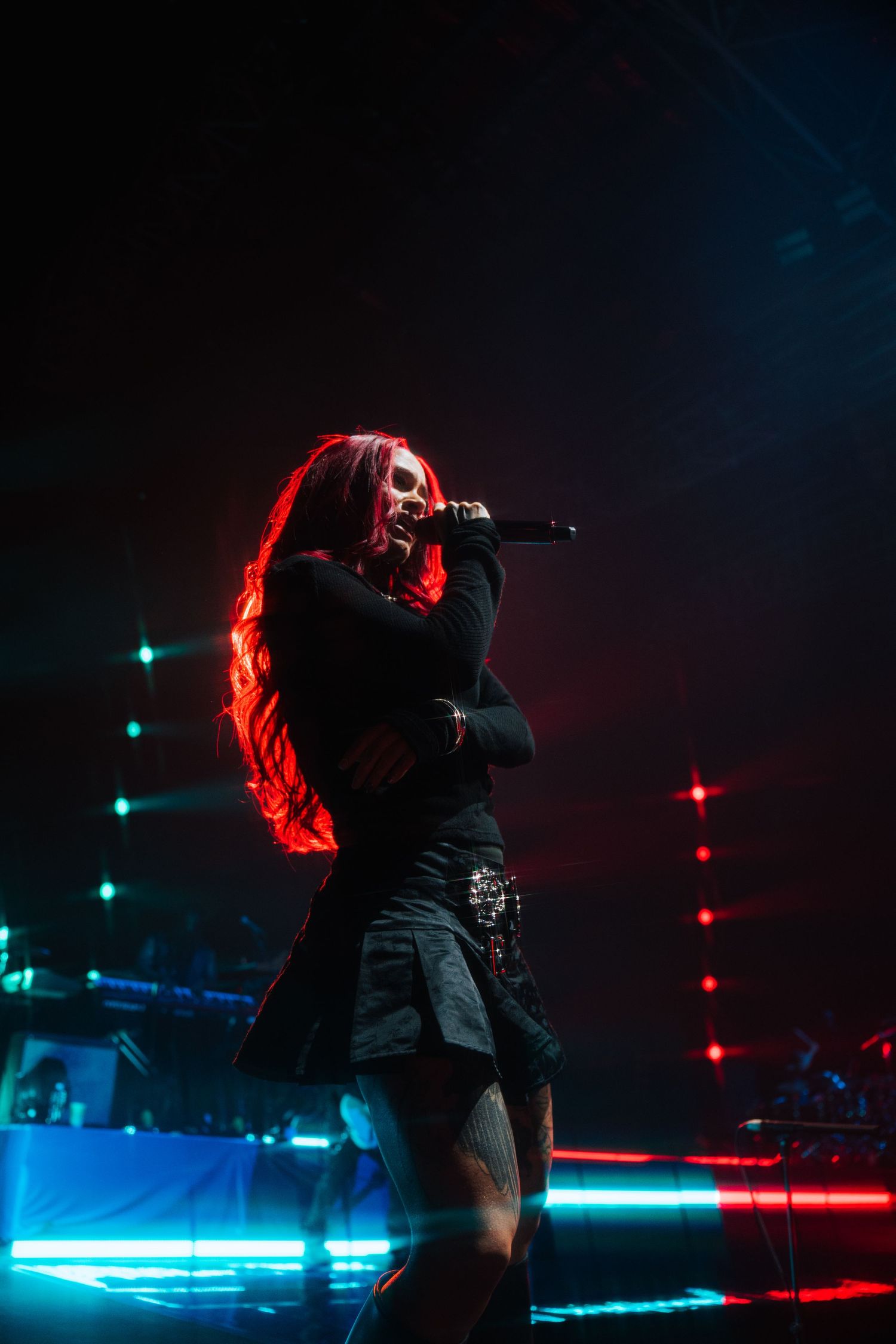 Singer performs on stage bathed in dramatic red and blue lighting with long flowing hair and black outfit during concert.
