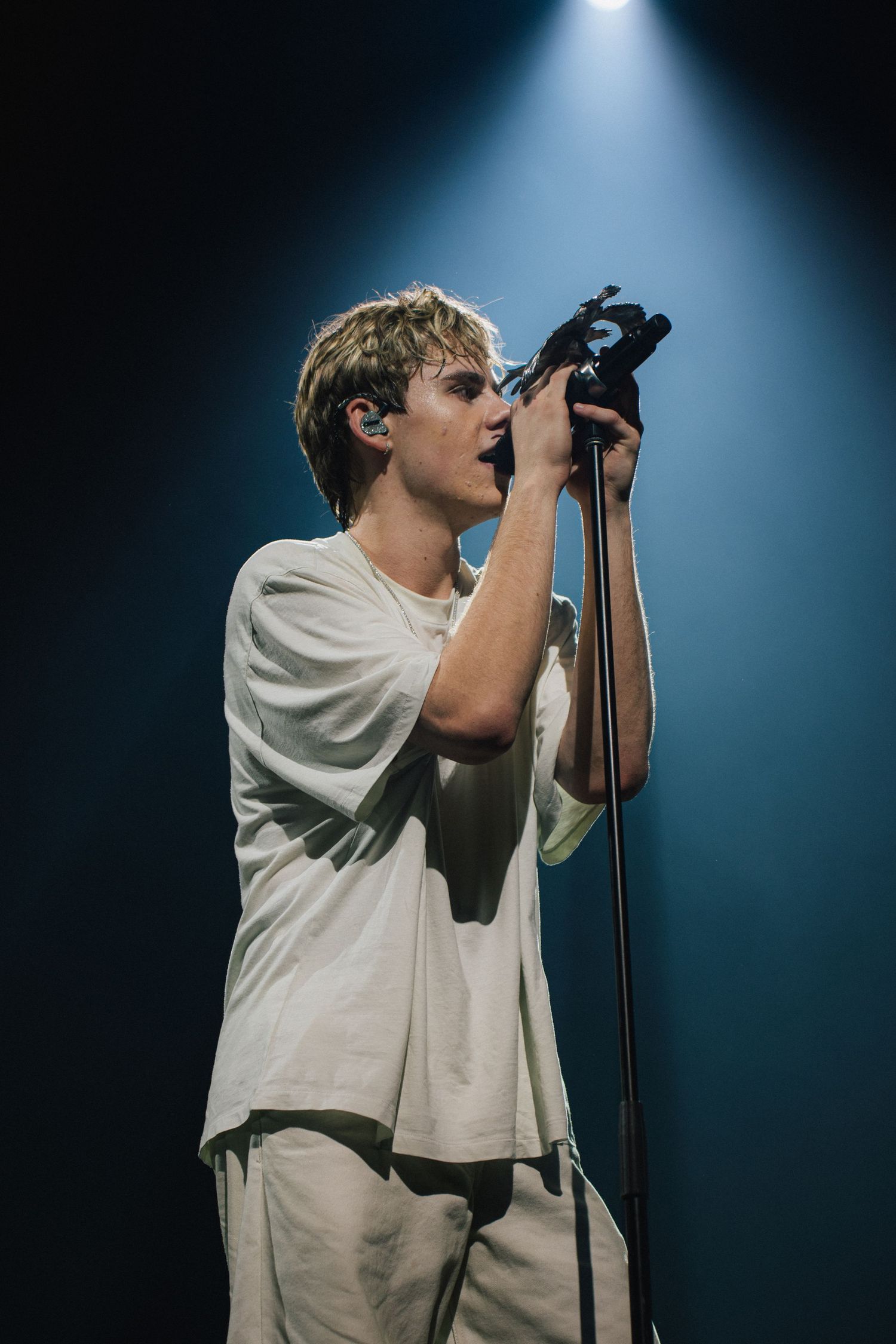 Singer in a white outfit performs on stage under dramatic blue spotlight during live concert performance.