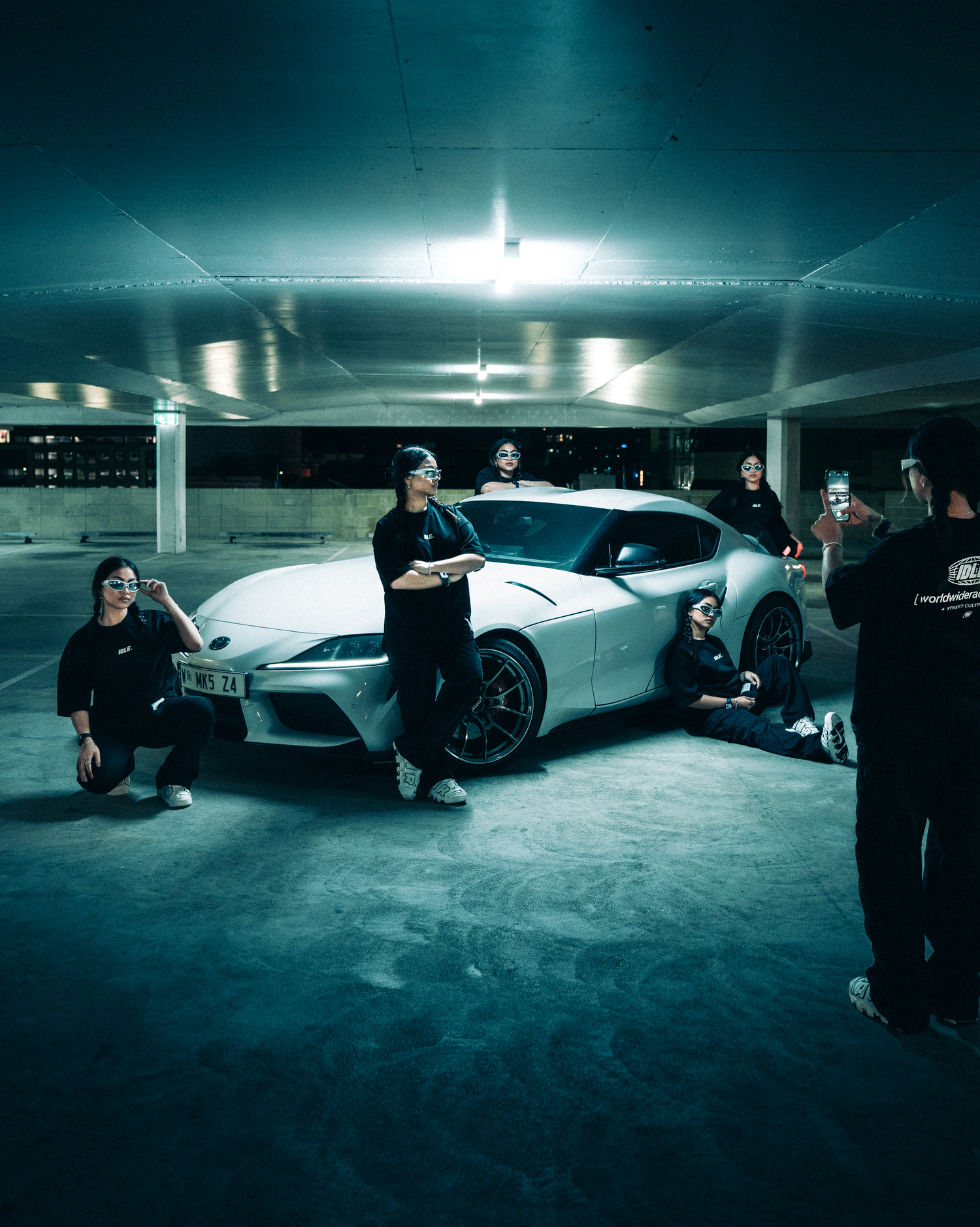 Group poses with sleek white sports car in dimly lit underground parking garage.