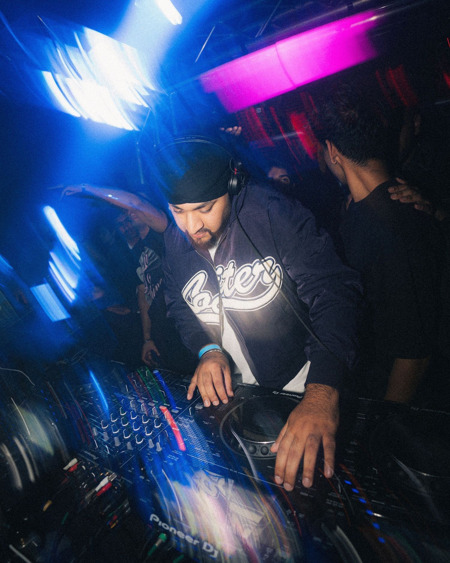 DJ in black hoodie mixing on turntables under blue and pink stage lighting.