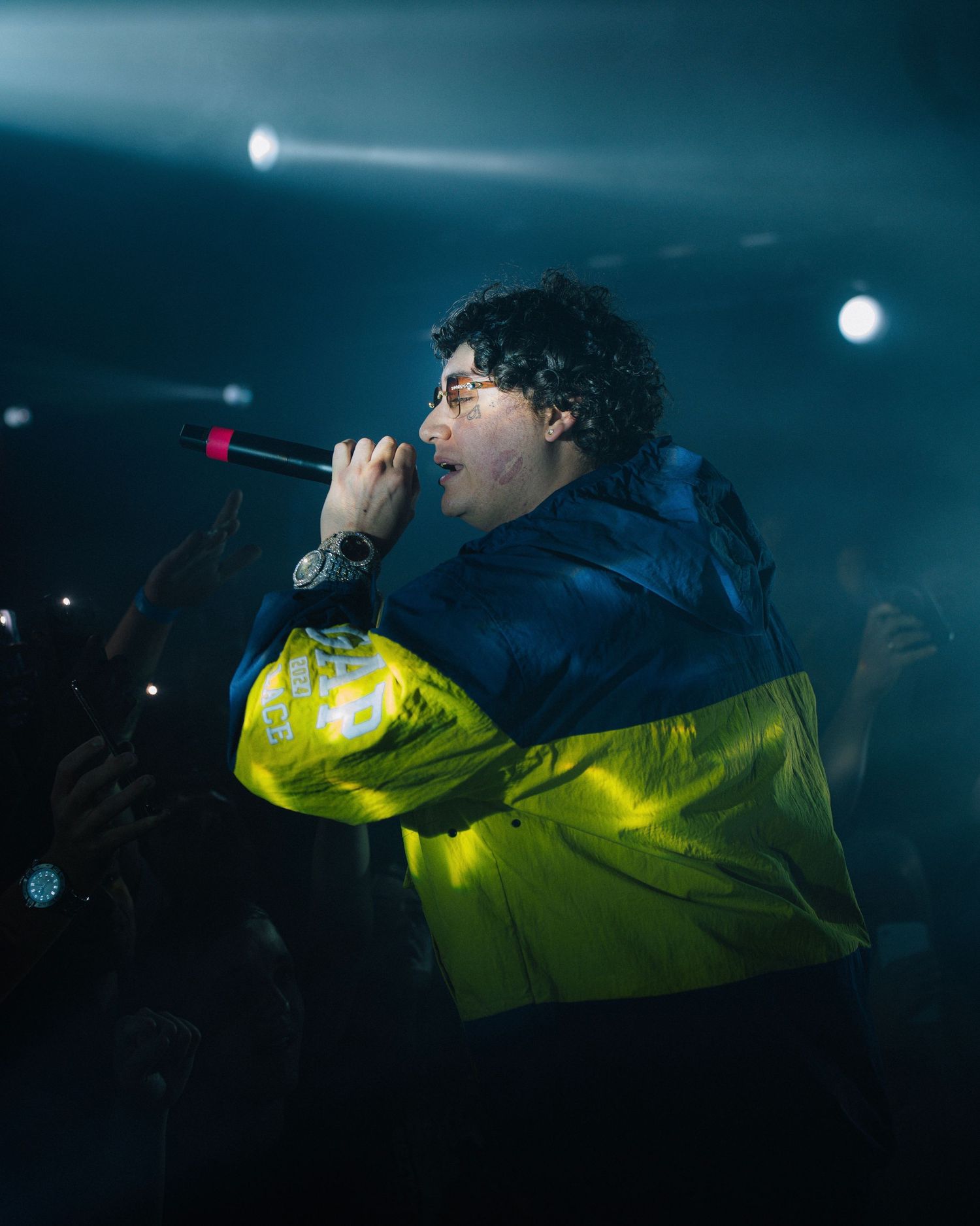 Musical artist performing in yellow and blue jacket under dramatic stage lighting.