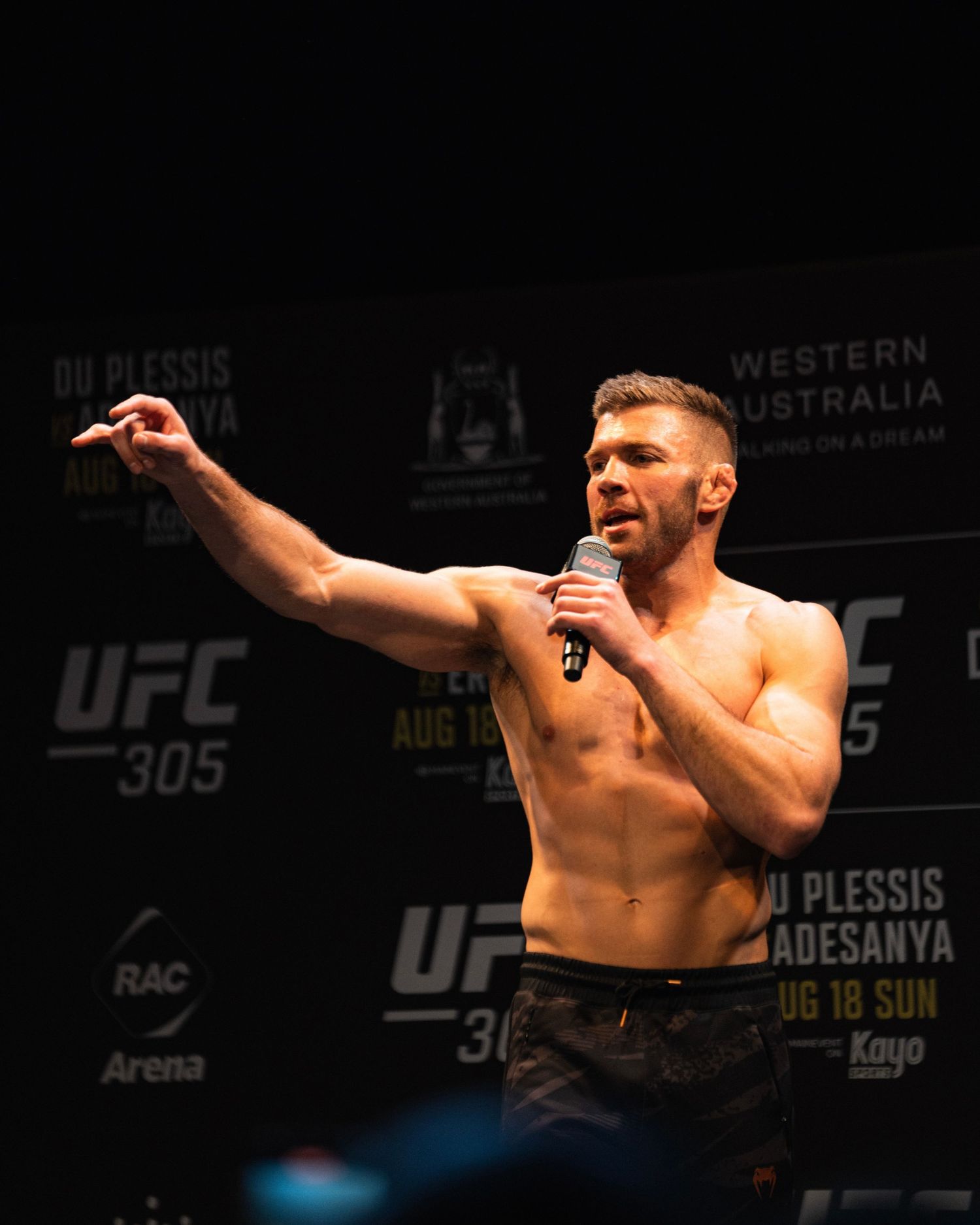 Shirtless UFC fighter gesturing while speaking at pre-fight event with Western Australia backdrop.