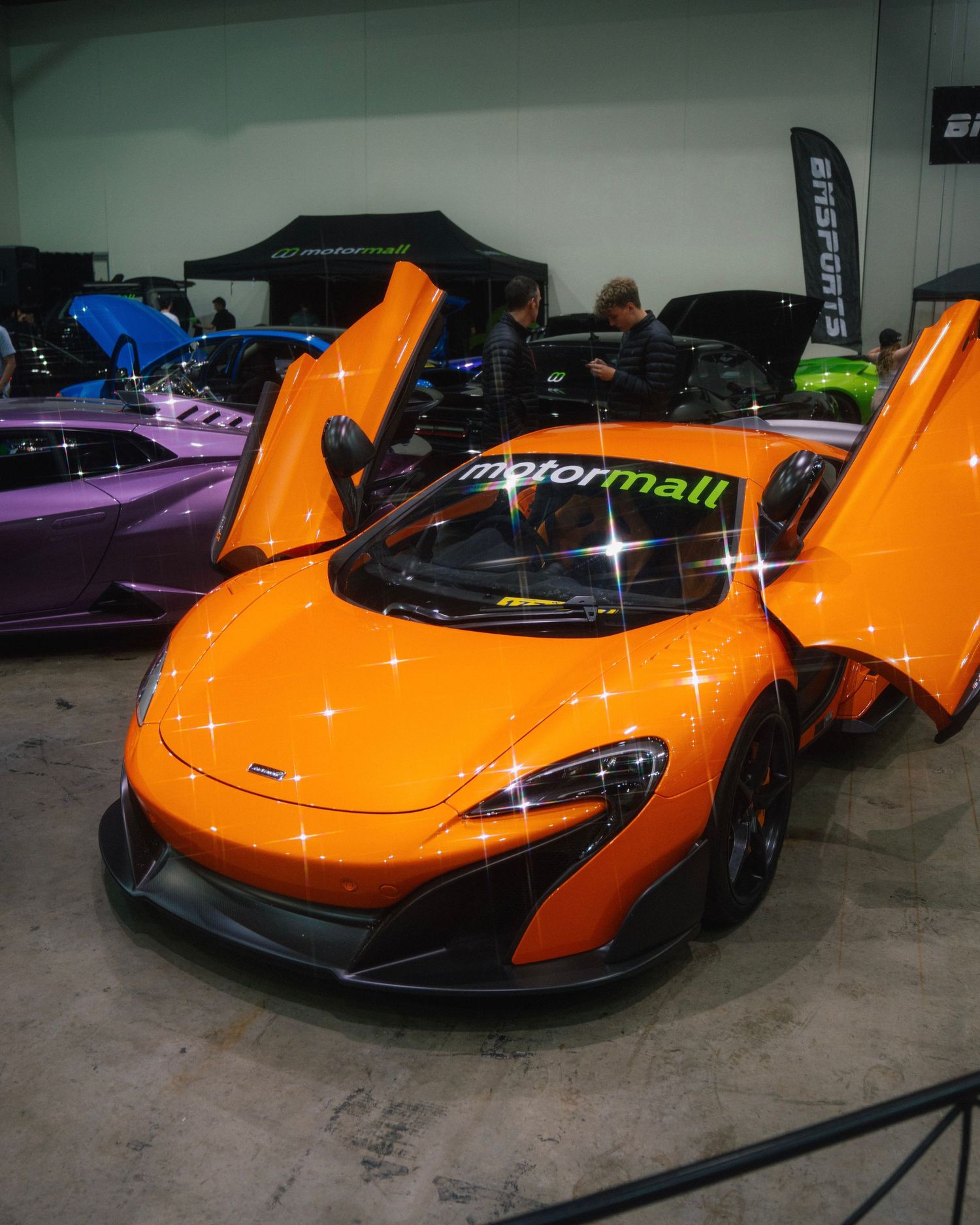 Orange McLaren supercar with butterfly doors open displayed at indoor car show.