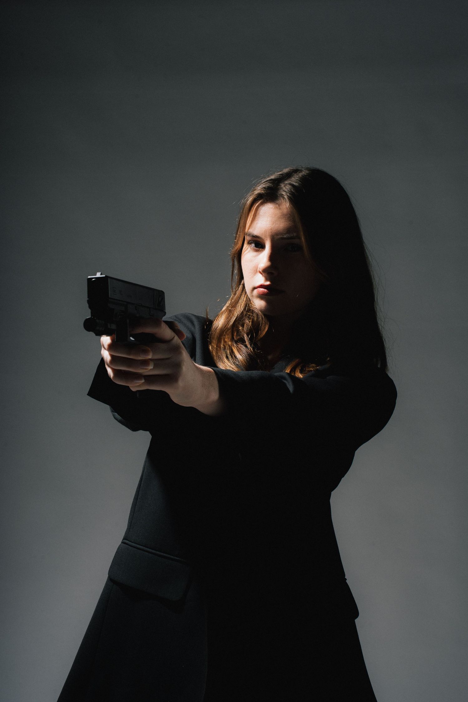 Person in black attire holding a handgun in a defensive stance against gray background.