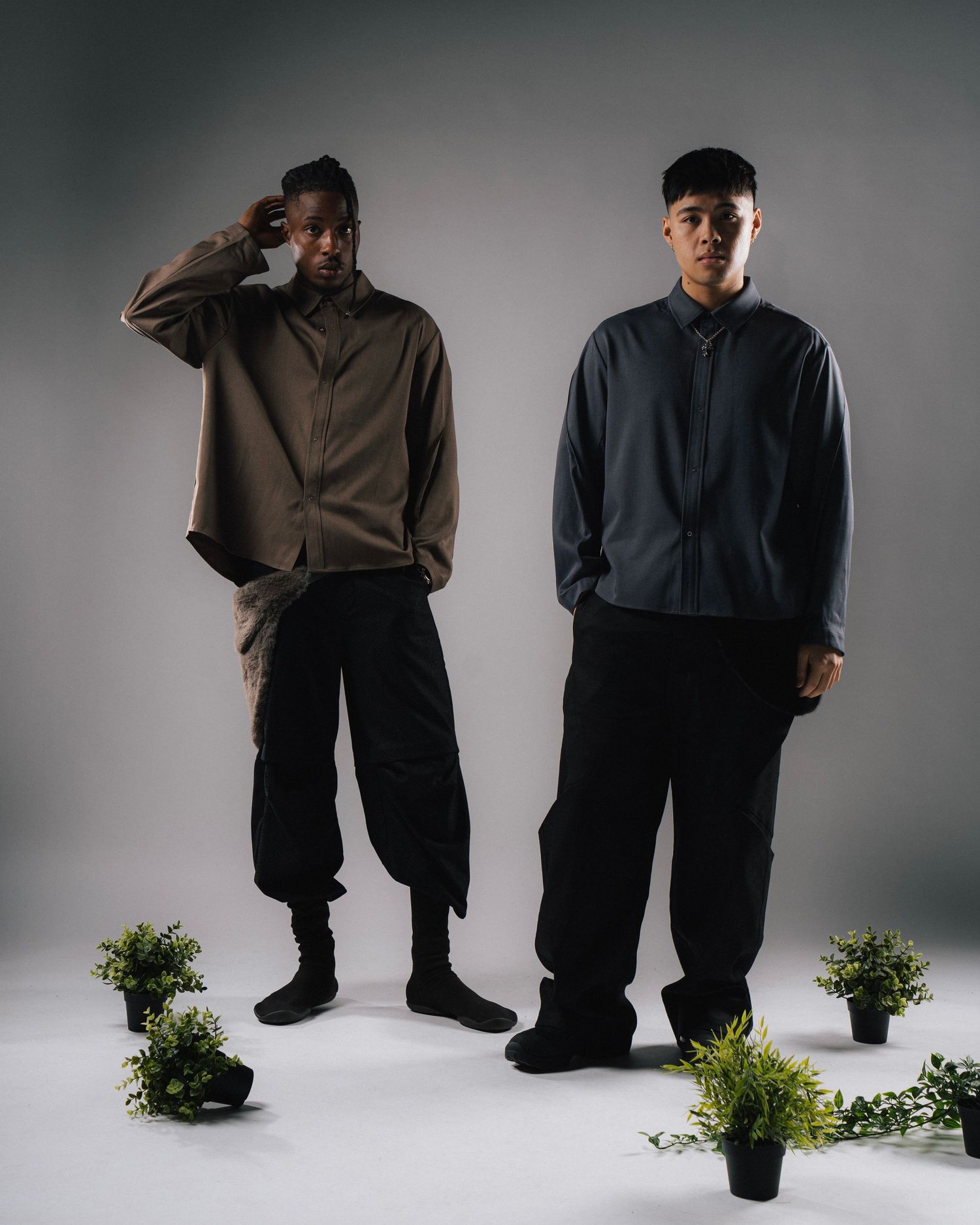 Two models in loose fitting streetwear standing with potted plants in studio setting.