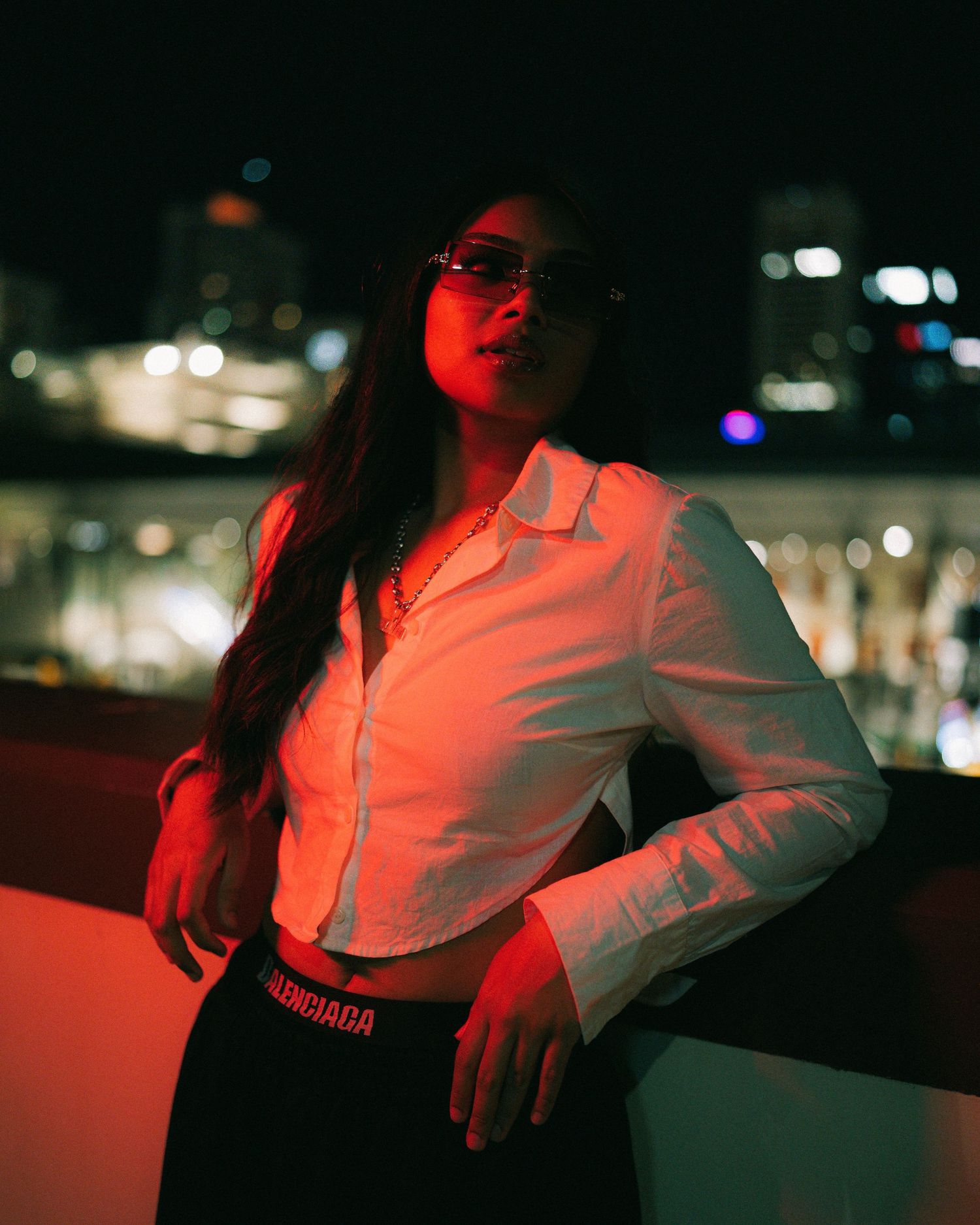 Woman in white shirt and sunglasses poses against city lights at night with red ambient lighting.