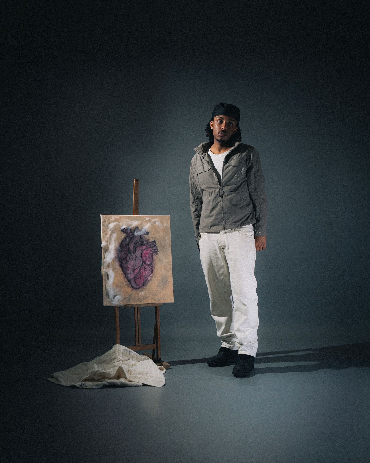 Artist stands next to an easel displaying an anatomical heart painting in a dramatic studio setting with moody lighting.