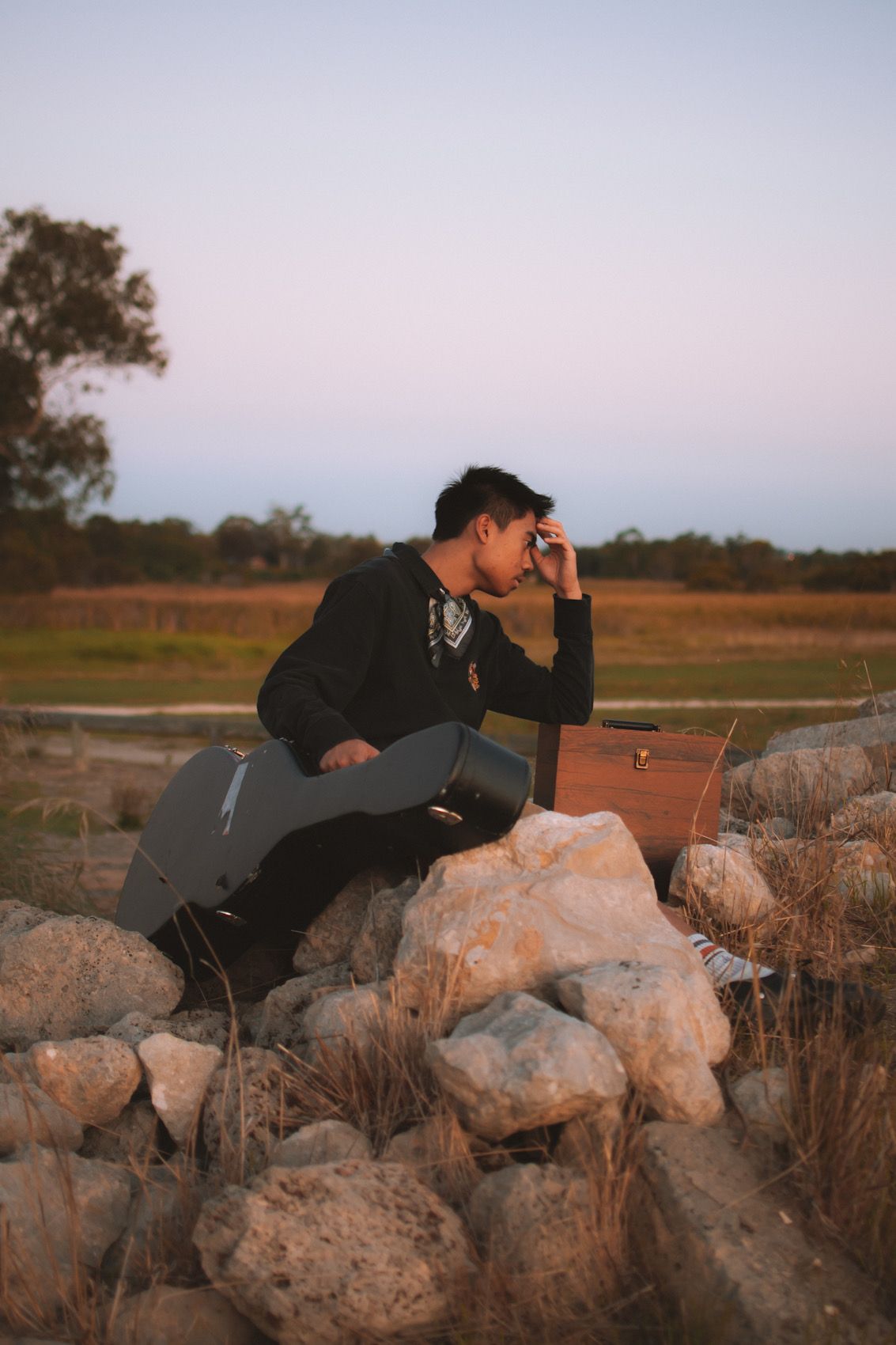 A person in dark clothing sits pensively on large rocks at sunset in a rural landscape.