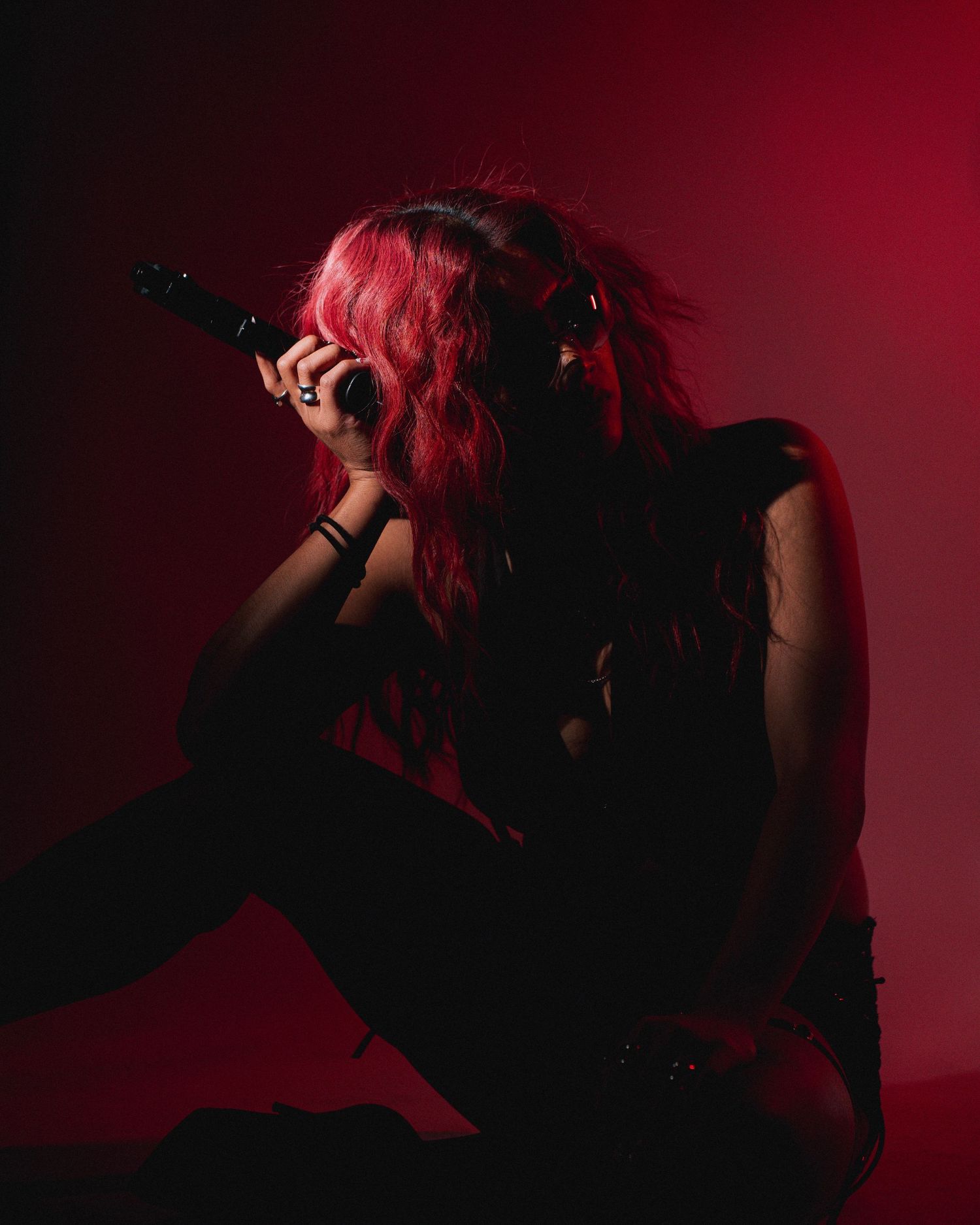 Figure in black clothing with vibrant pink hair poses dramatically against dark red background in moody portrait.