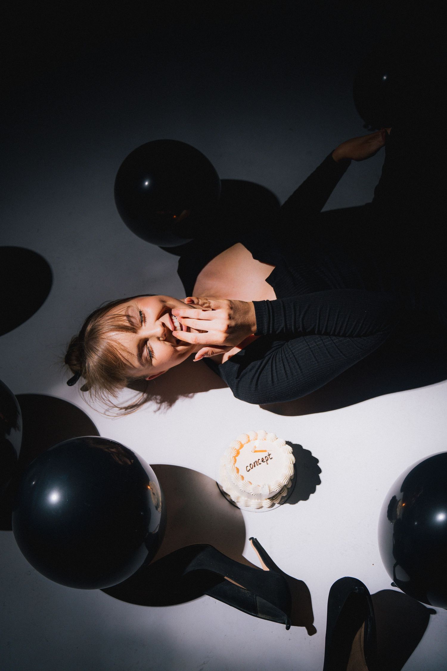 An artistic overhead photo with black balloons and white background featuring someone in dark clothing laying down.