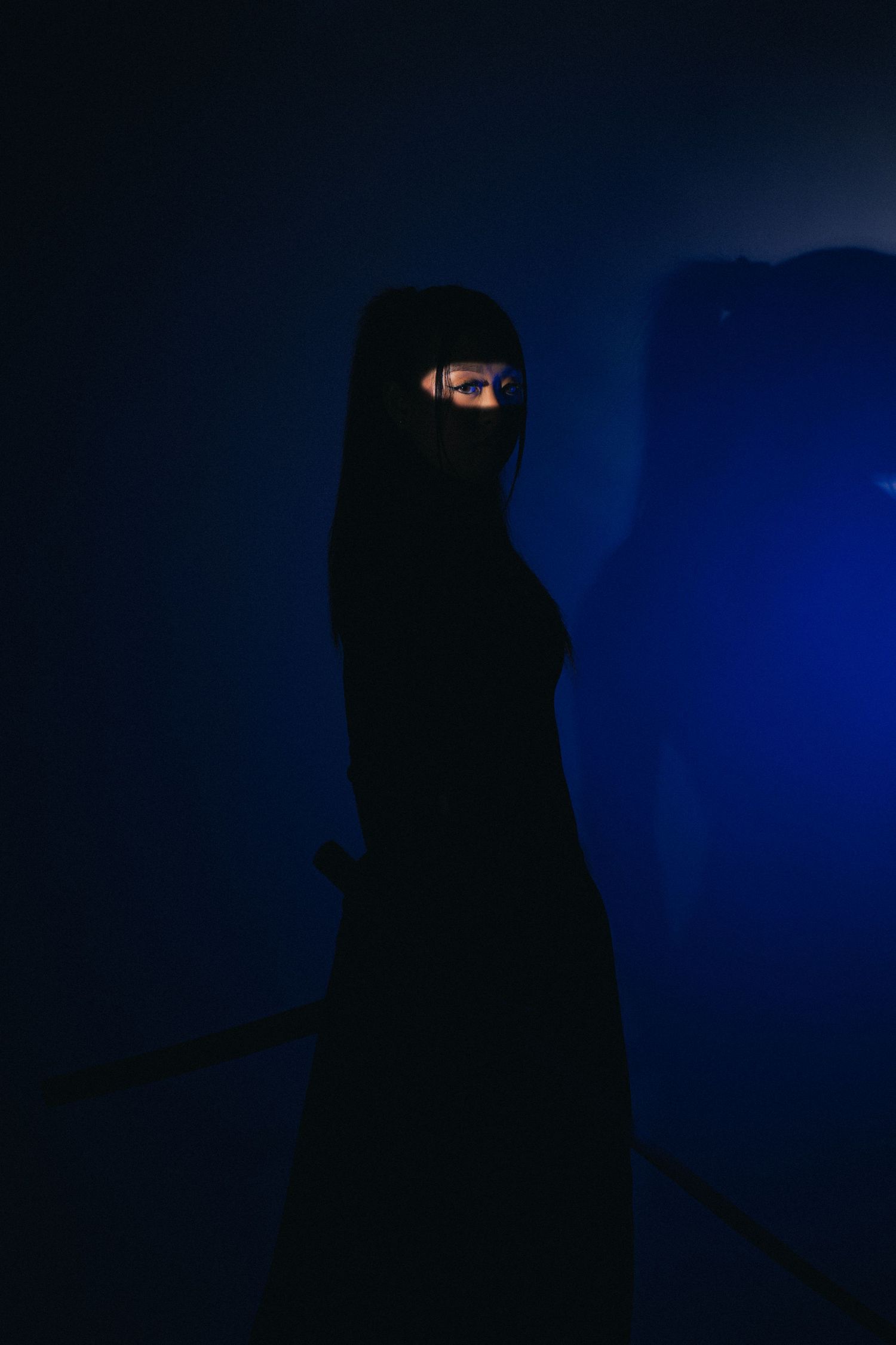Figure in dark abaya stands against dramatic blue backdrop in moody low-light portrait photography.