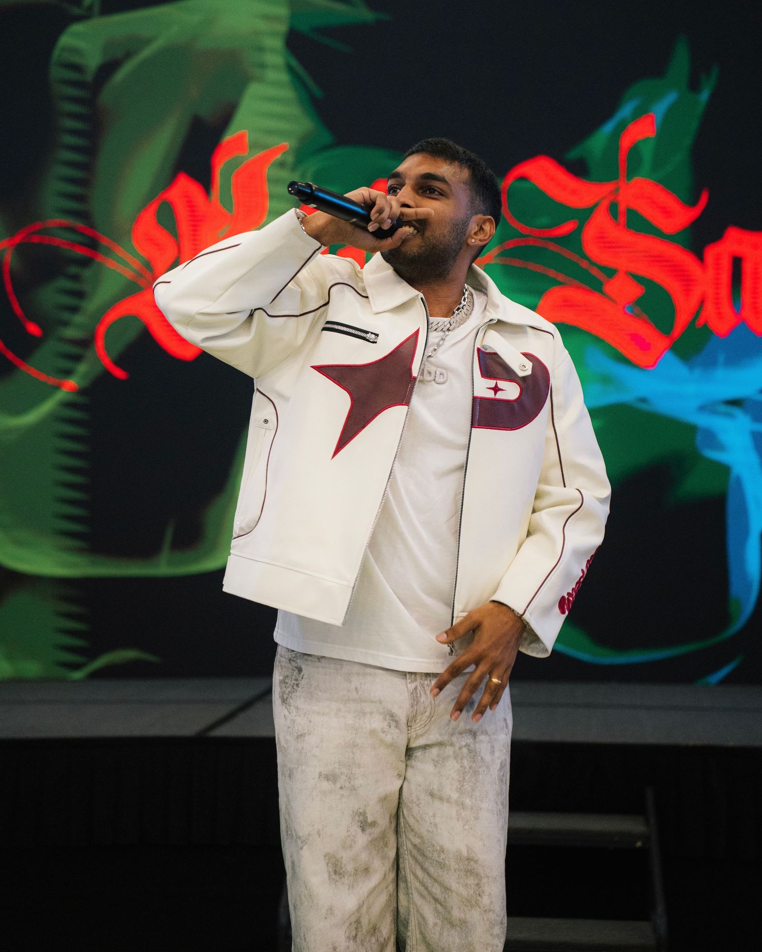 A performer in a white jacket with burgundy accents sings into a microphone on stage with colorful lights in the background.
