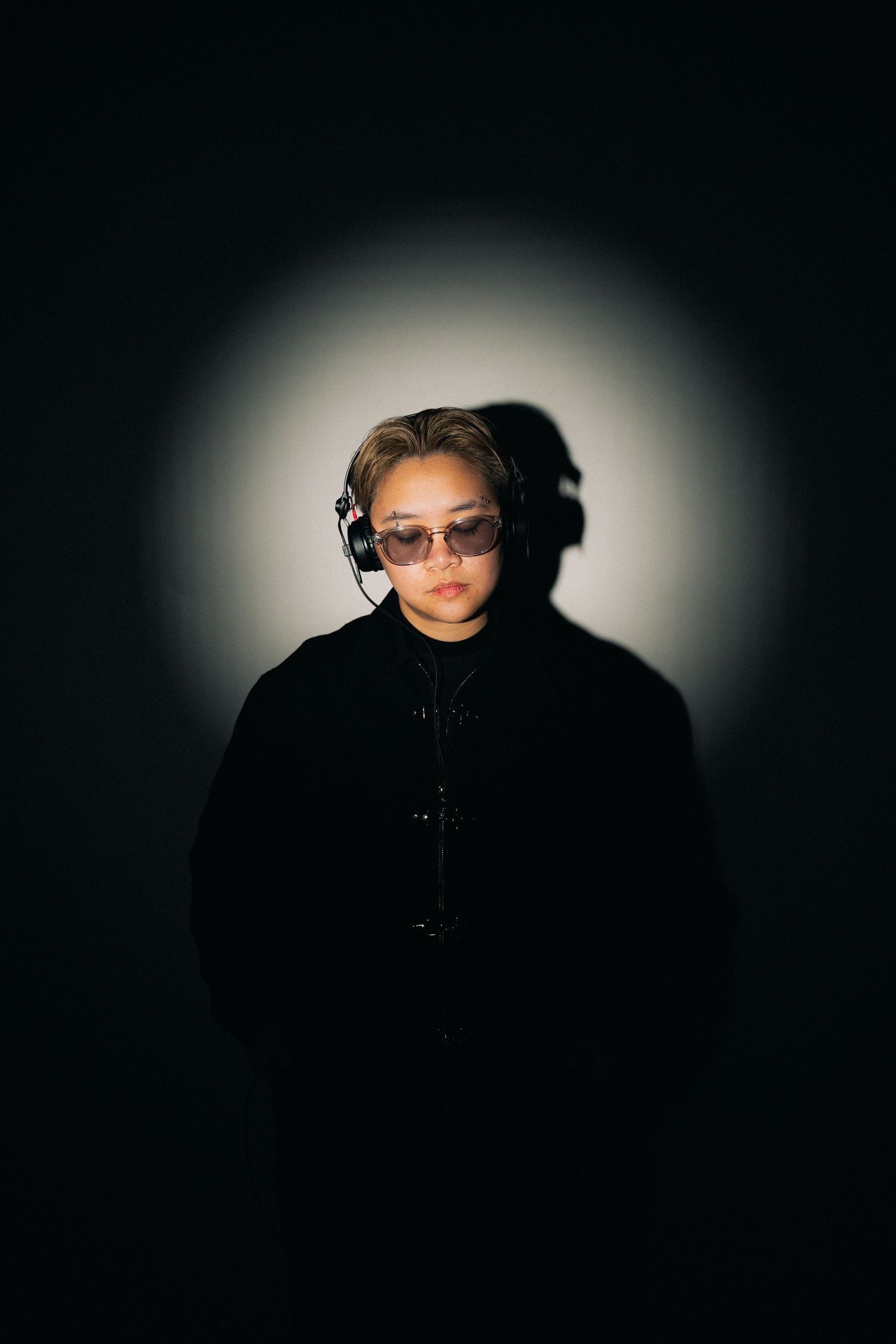 Figure wearing sunglasses and headphones photographed against dark background with dramatic halo lighting effect.