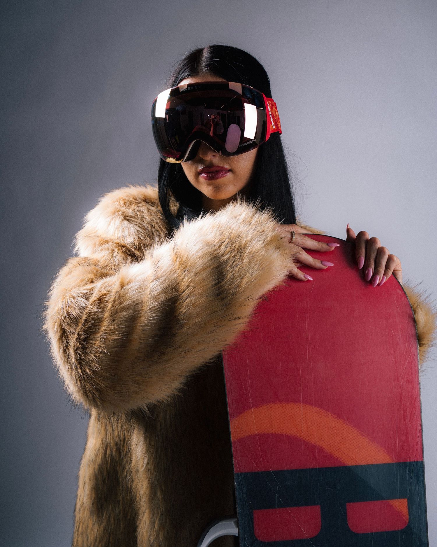Snowboarder in a fur coat and VR goggles holds a red snowboard against a gray studio background.