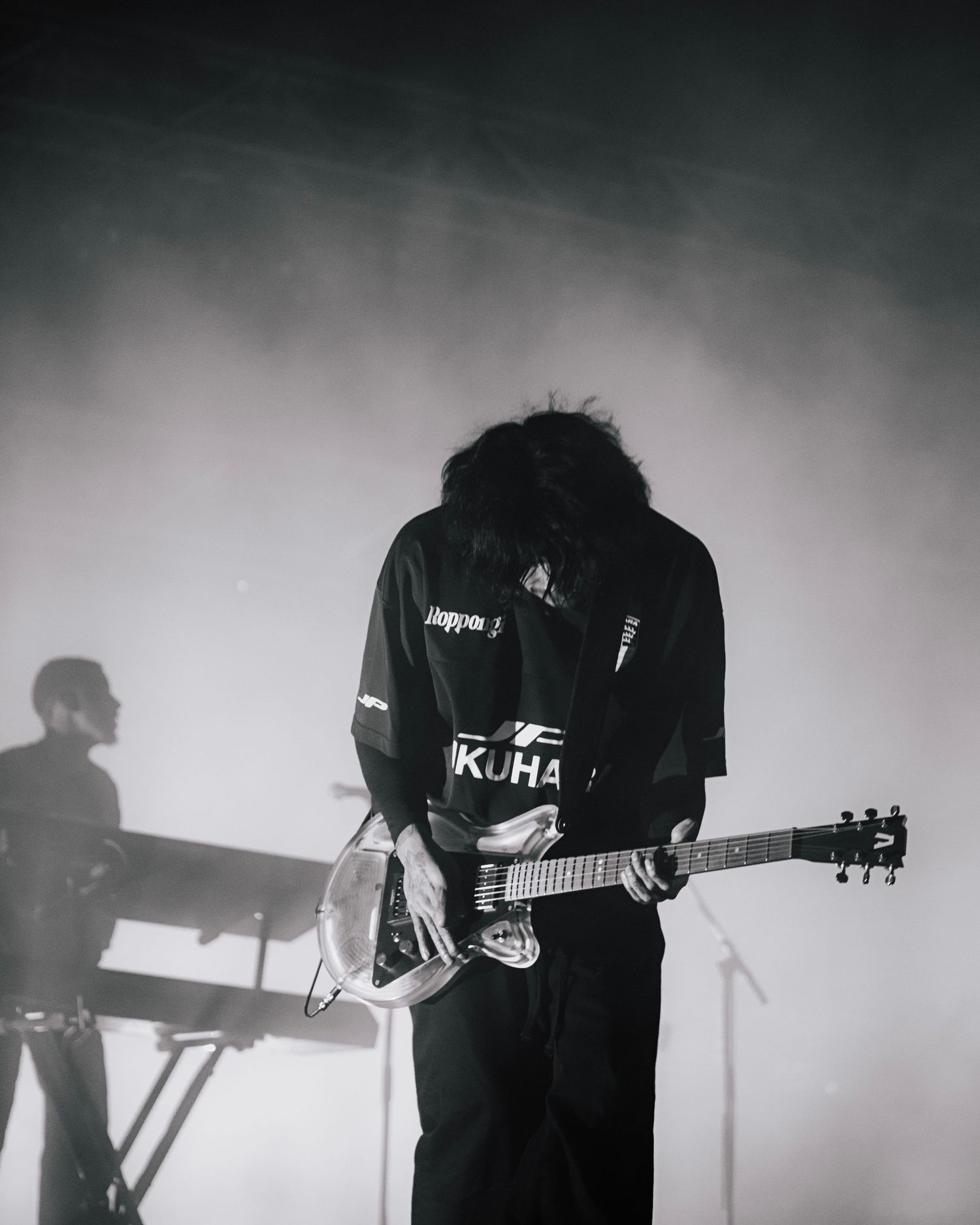 A guitarist in dark clothing performs on stage with a silhouetted keyboard player visible in the atmospheric background.