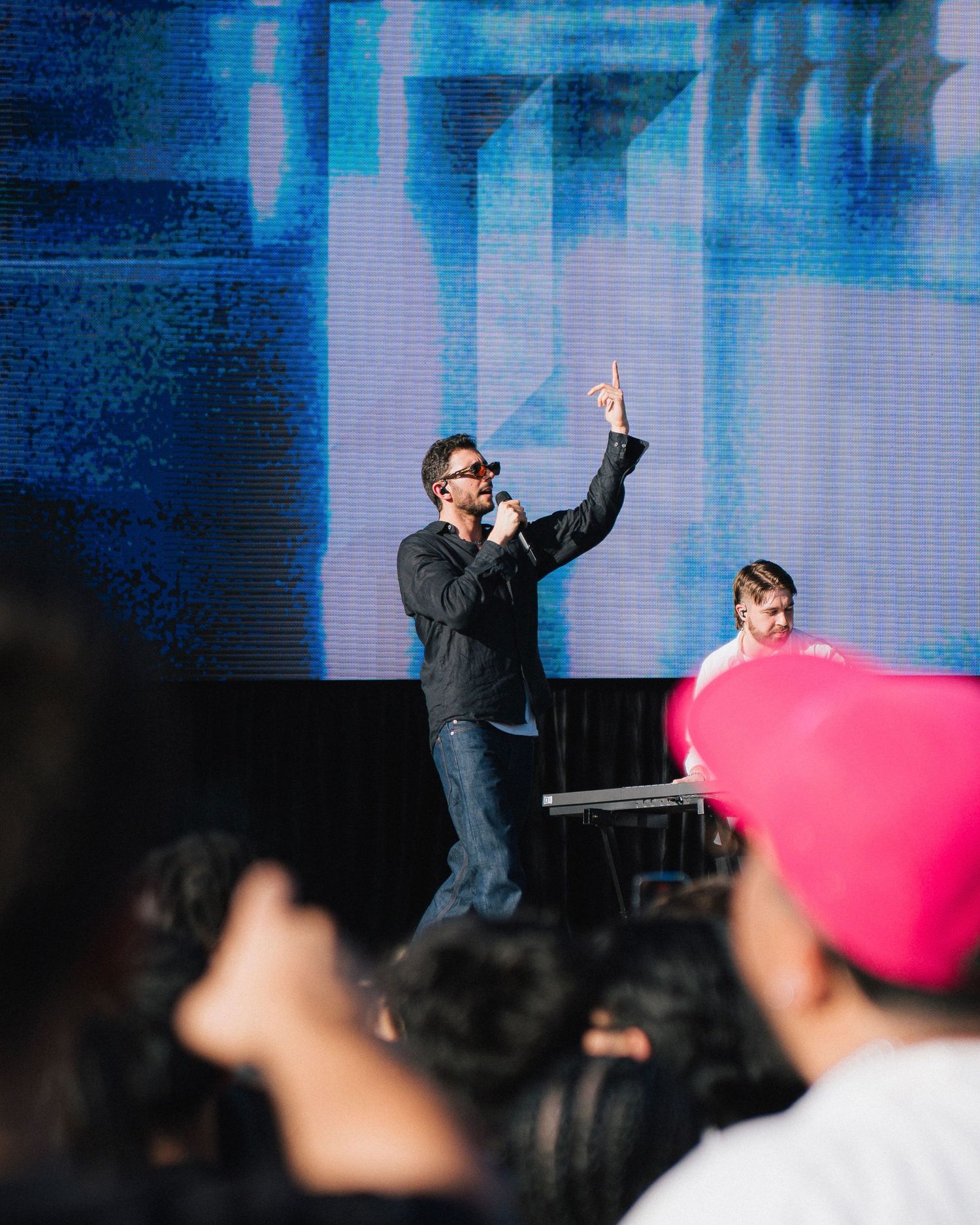 A performer in dark attire raises their hand while on stage with blue lighting and an audience in the foreground.
