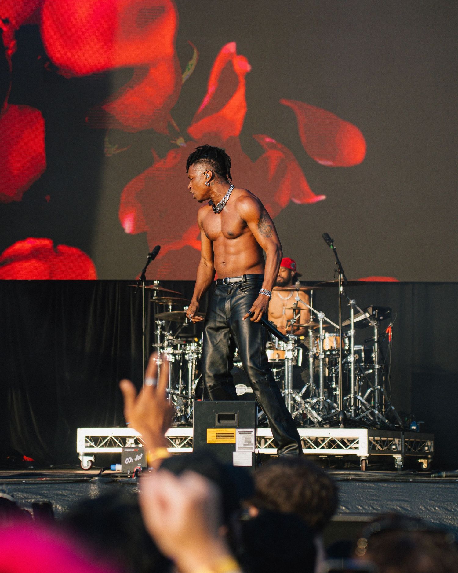 Shirtless performer on stage with a dramatic red rose backdrop and drum kit during an energetic concert performance.