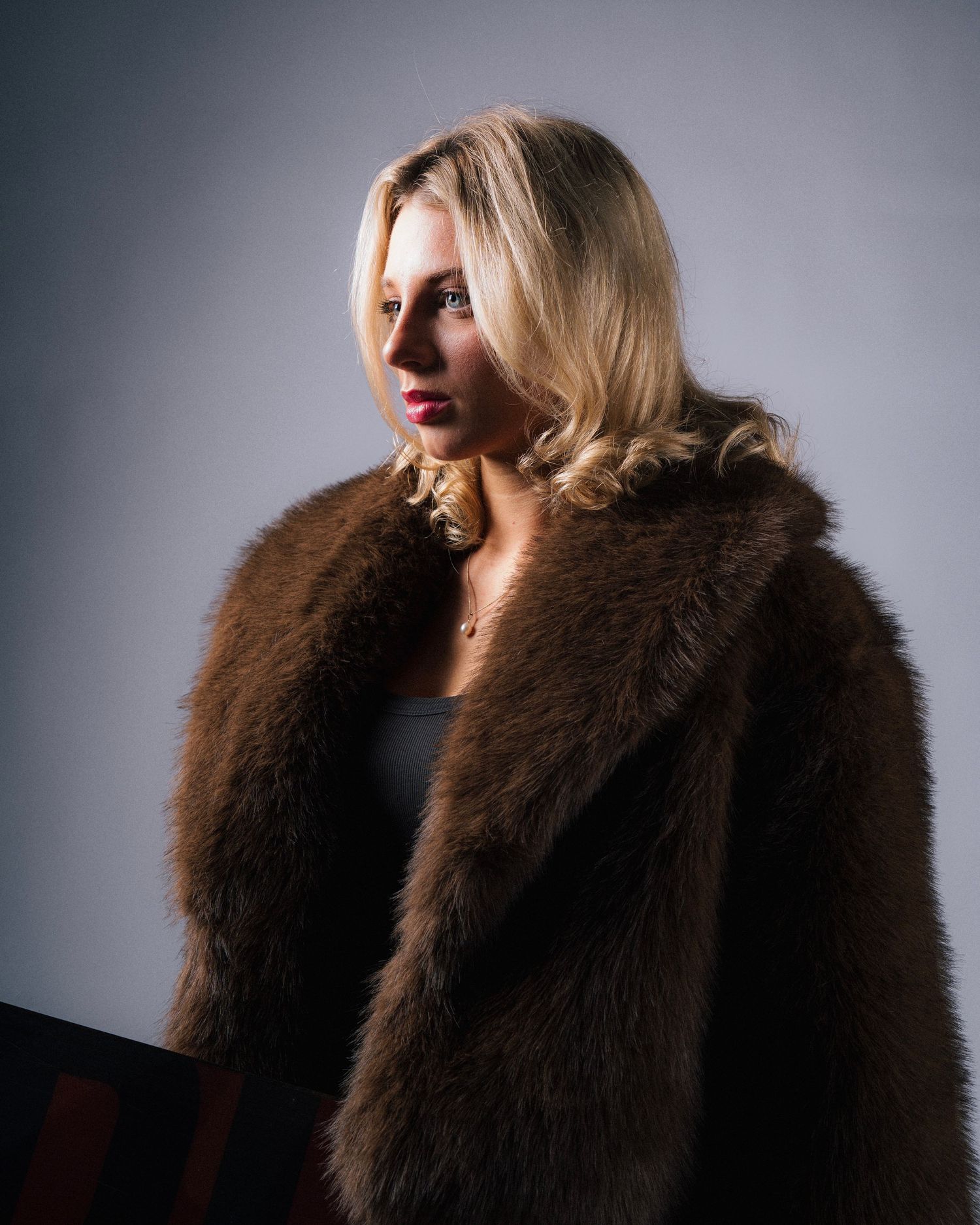 A woman wearing a dramatic dark brown fur coat poses against a grey studio backdrop in a glamorous fashion series.