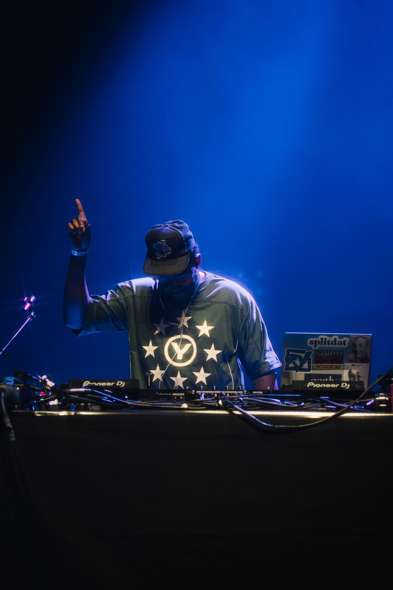 DJ performs on stage wearing a starred t-shirt while mixing music on turntables under dramatic blue lighting.