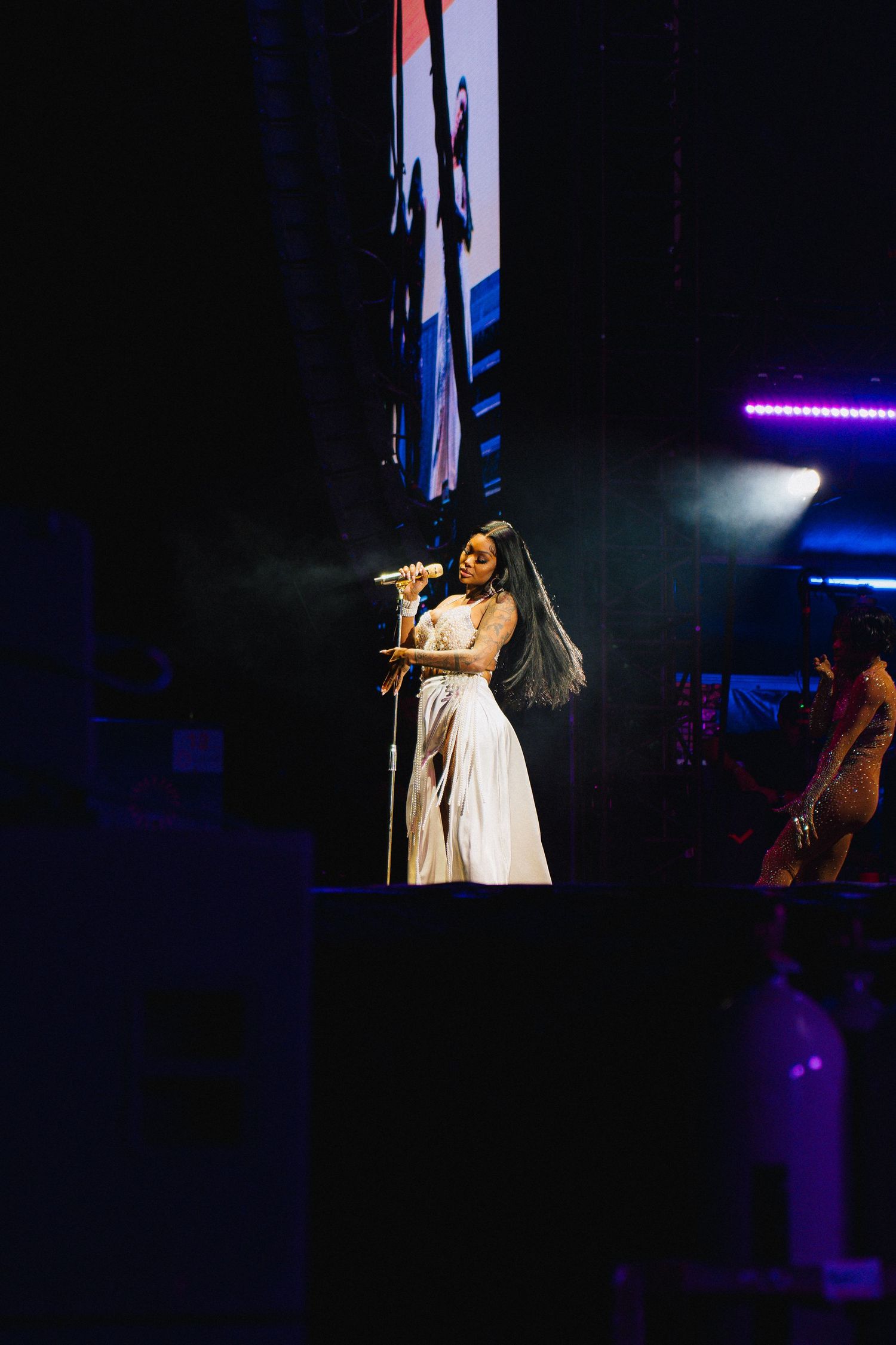 A performer in a white dress sings on a dark stage with purple lighting and screens in the background.