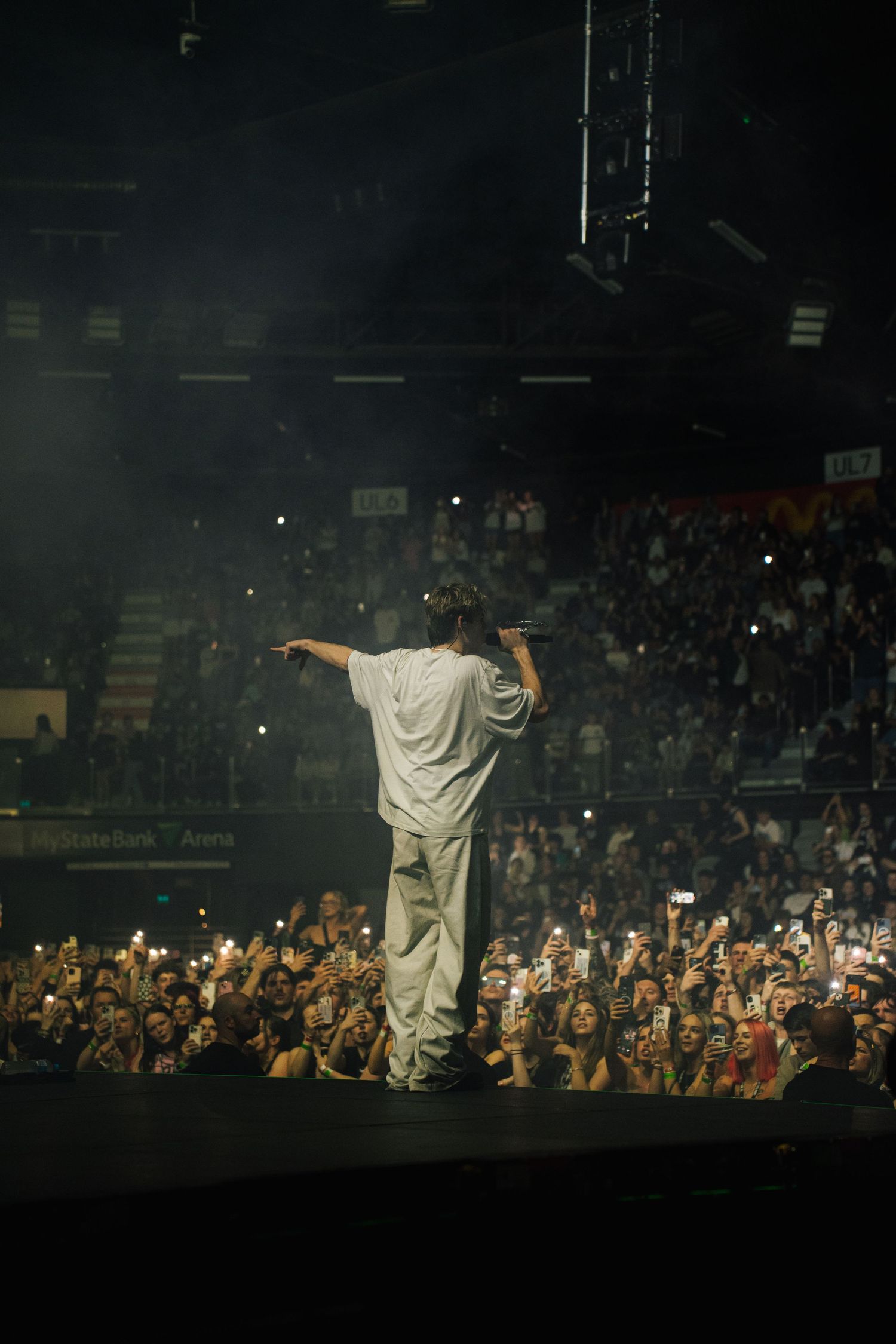 Performer in white stands silhouetted on dark stage before large crowd during concert performance.
