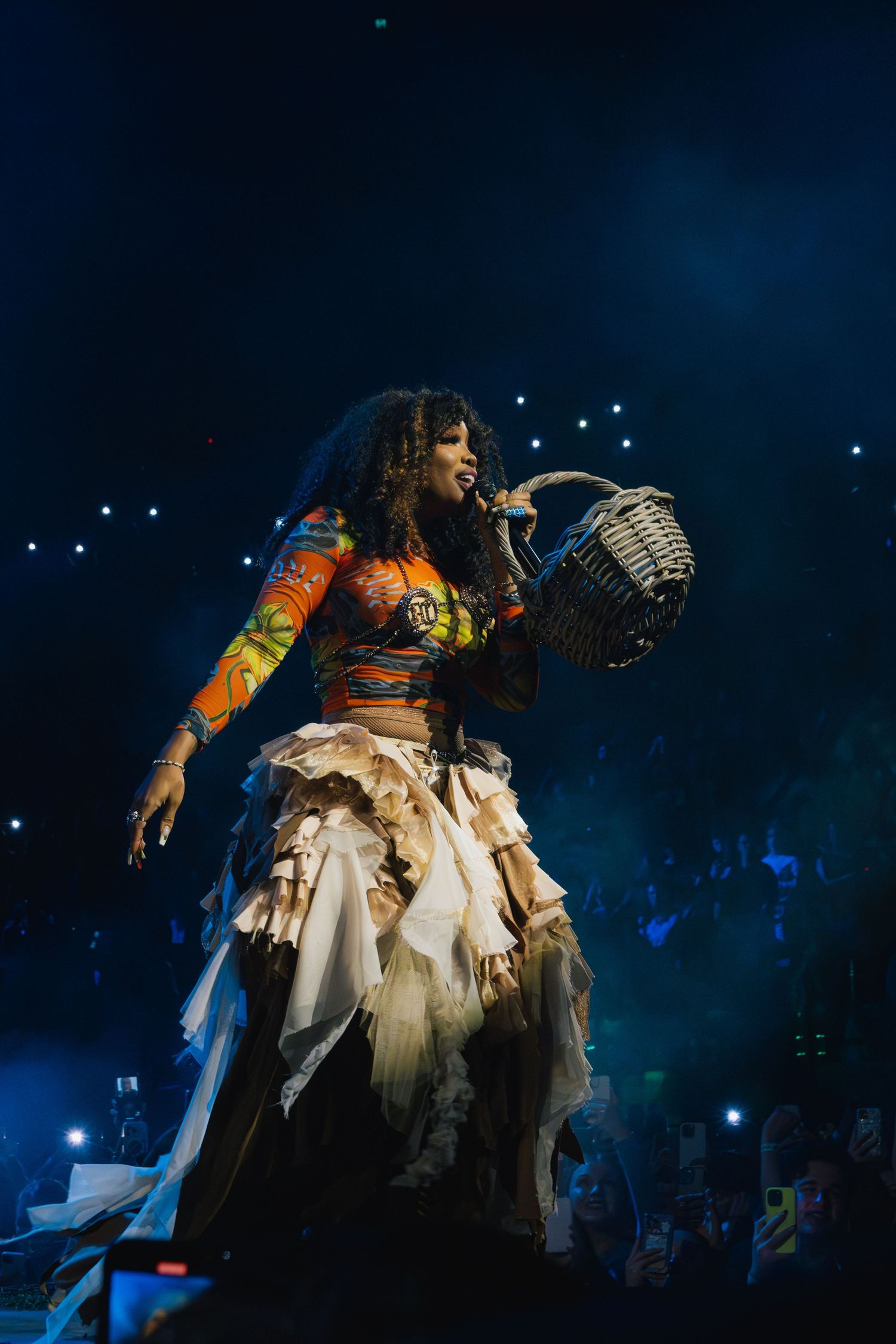 A performer in a colorful ruffled dress sings on stage during a dramatic concert performance with blue lighting.