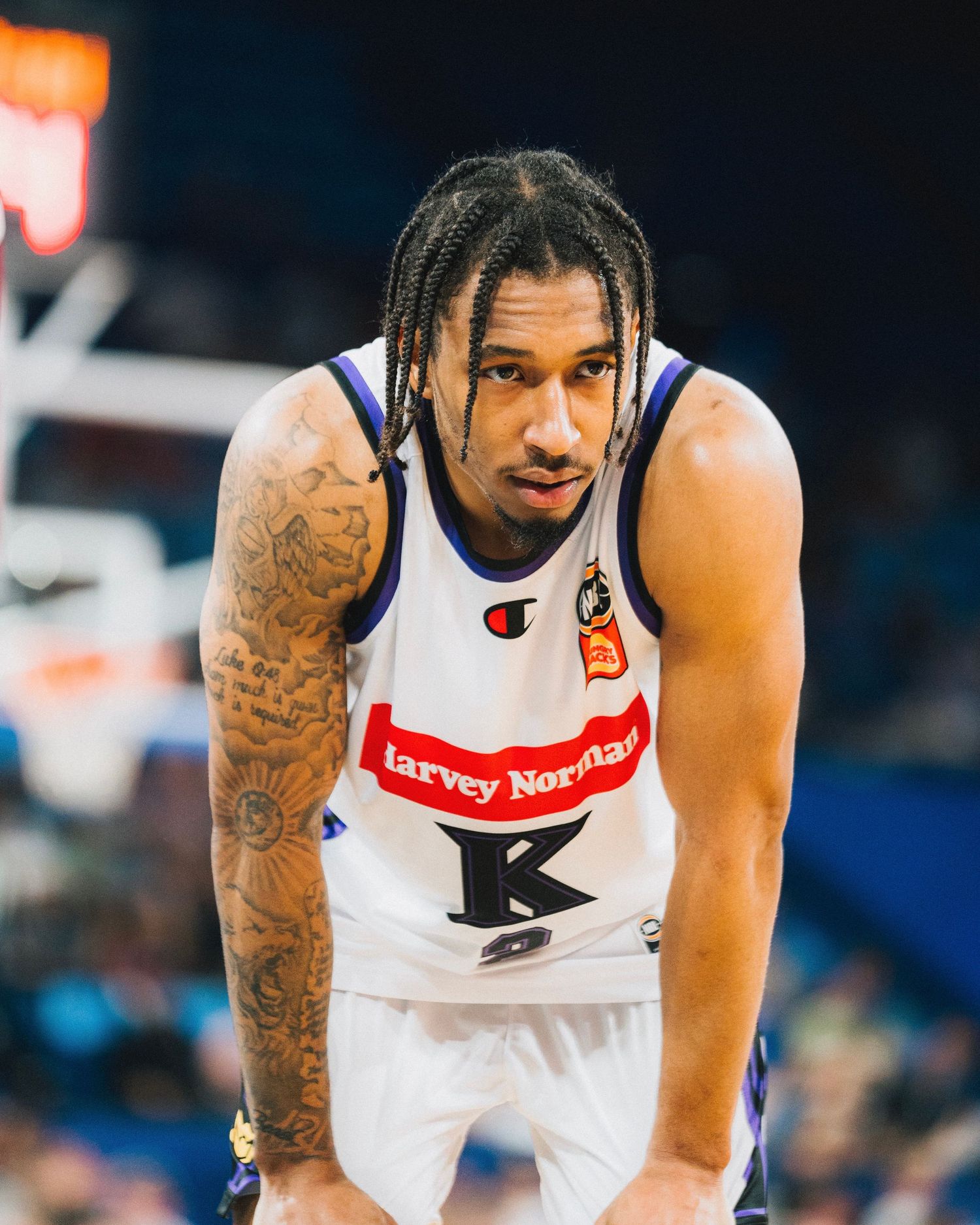 Basketball player in white jersey with tattoos and braids stands on court.