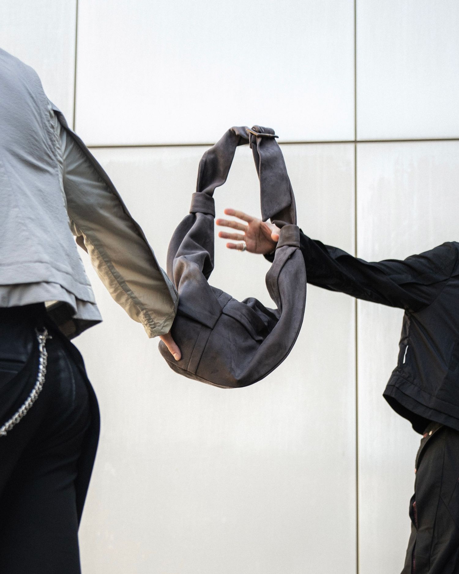 Two people passing a gray handbag between them against white tiled wall.