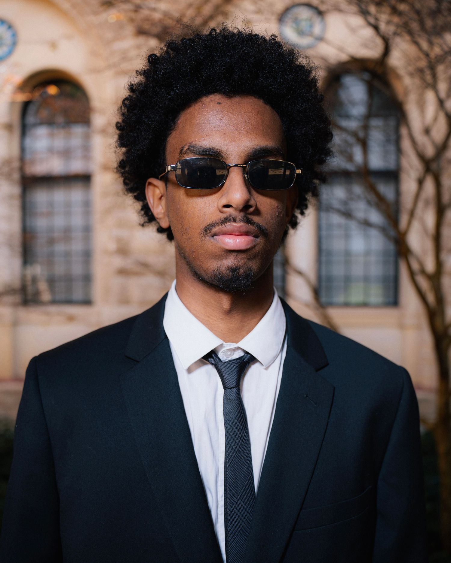 Professional portrait of a person in suit and sunglasses against architectural backdrop.