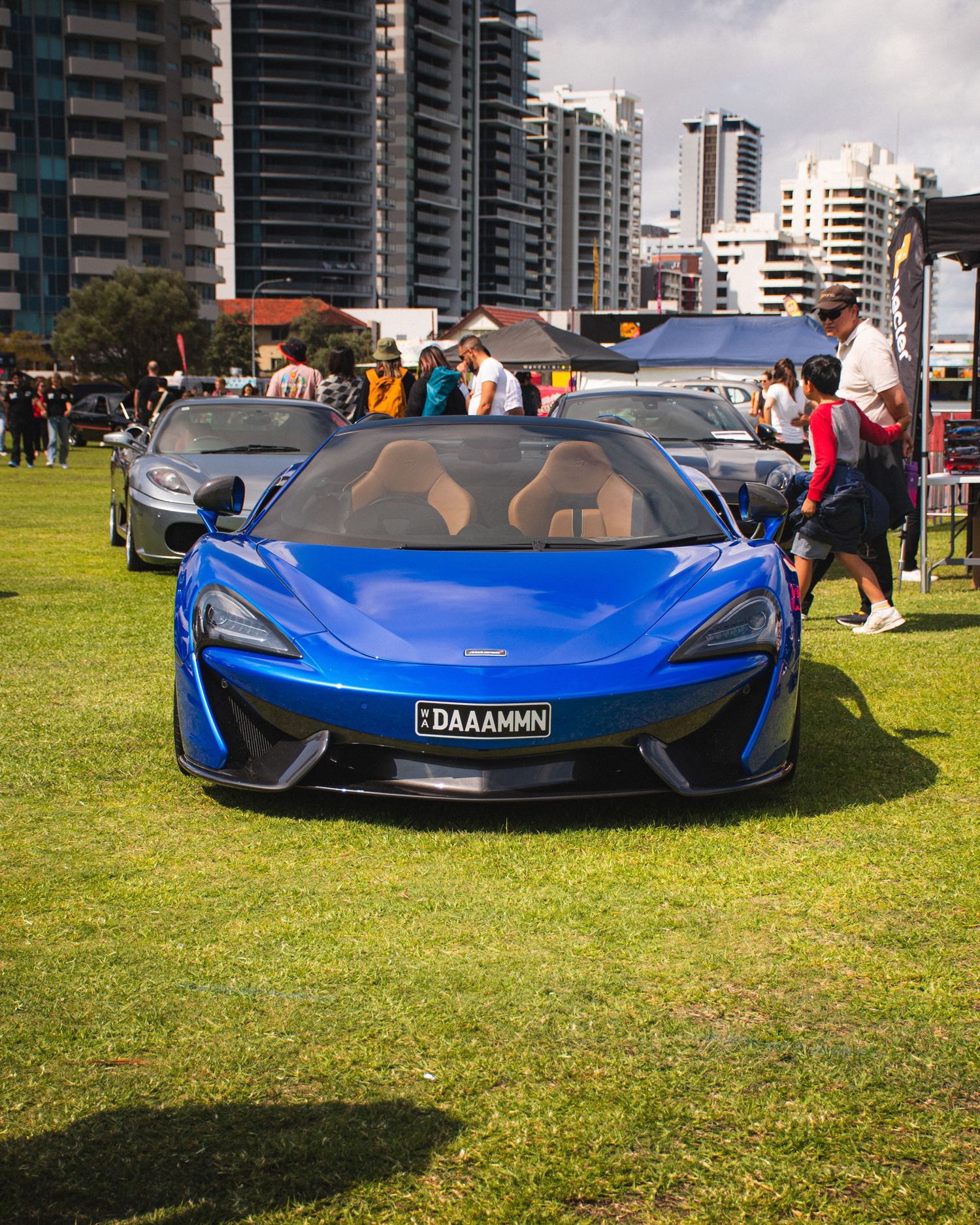Blue McLaren sports car displayed on grass at outdoor car show with high-rise buildings behind.