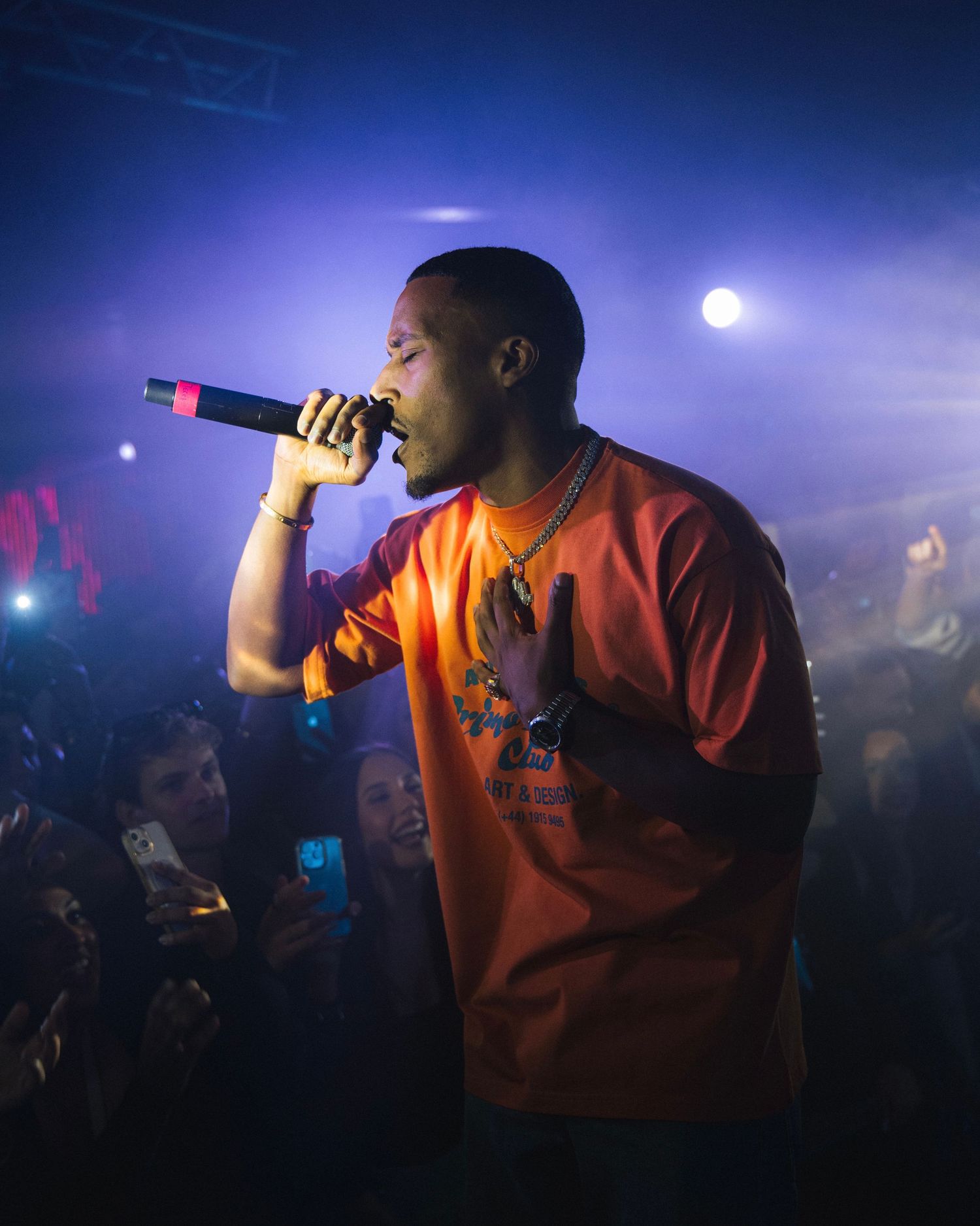 Performer in orange shirt holds microphone while performing at nightclub with purple lighting effects.