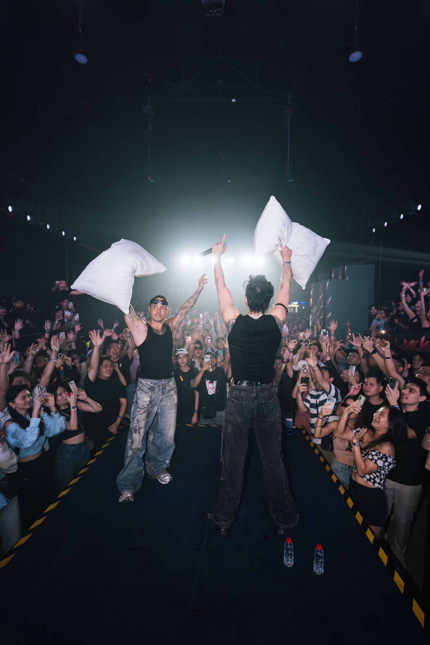 Two performers hold pillows aloft on stage as an excited crowd cheers during a high-energy concert performance.