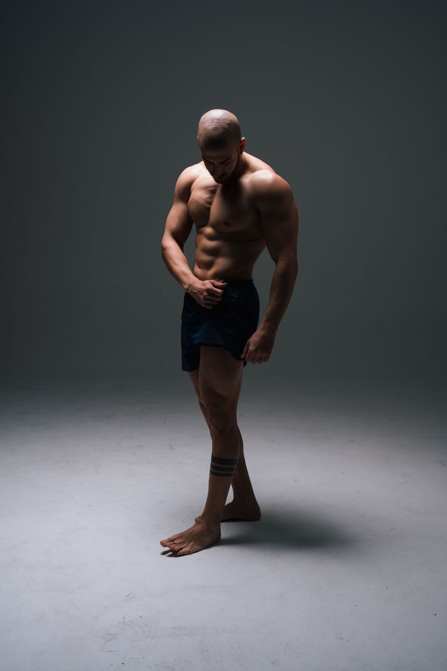 Athletic figure in black shorts poses against dark background showing muscular physique in dramatic studio lighting.