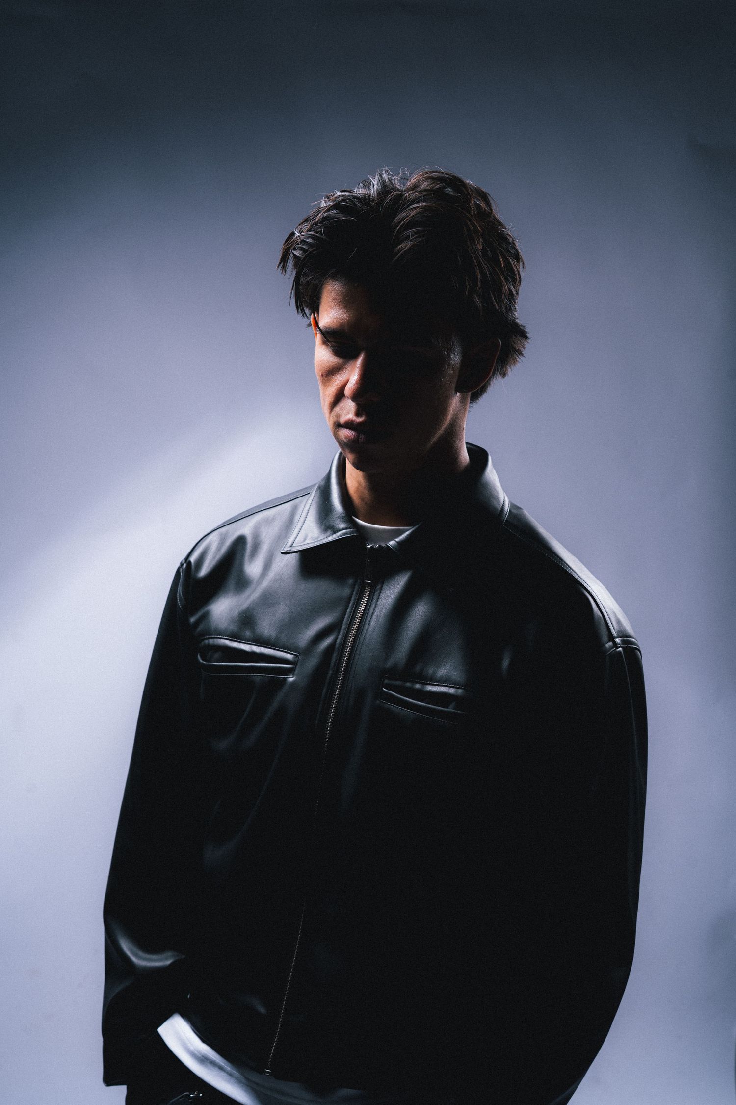 Dramatic studio portrait with moody lighting showing a person in a black leather jacket against a dark background.