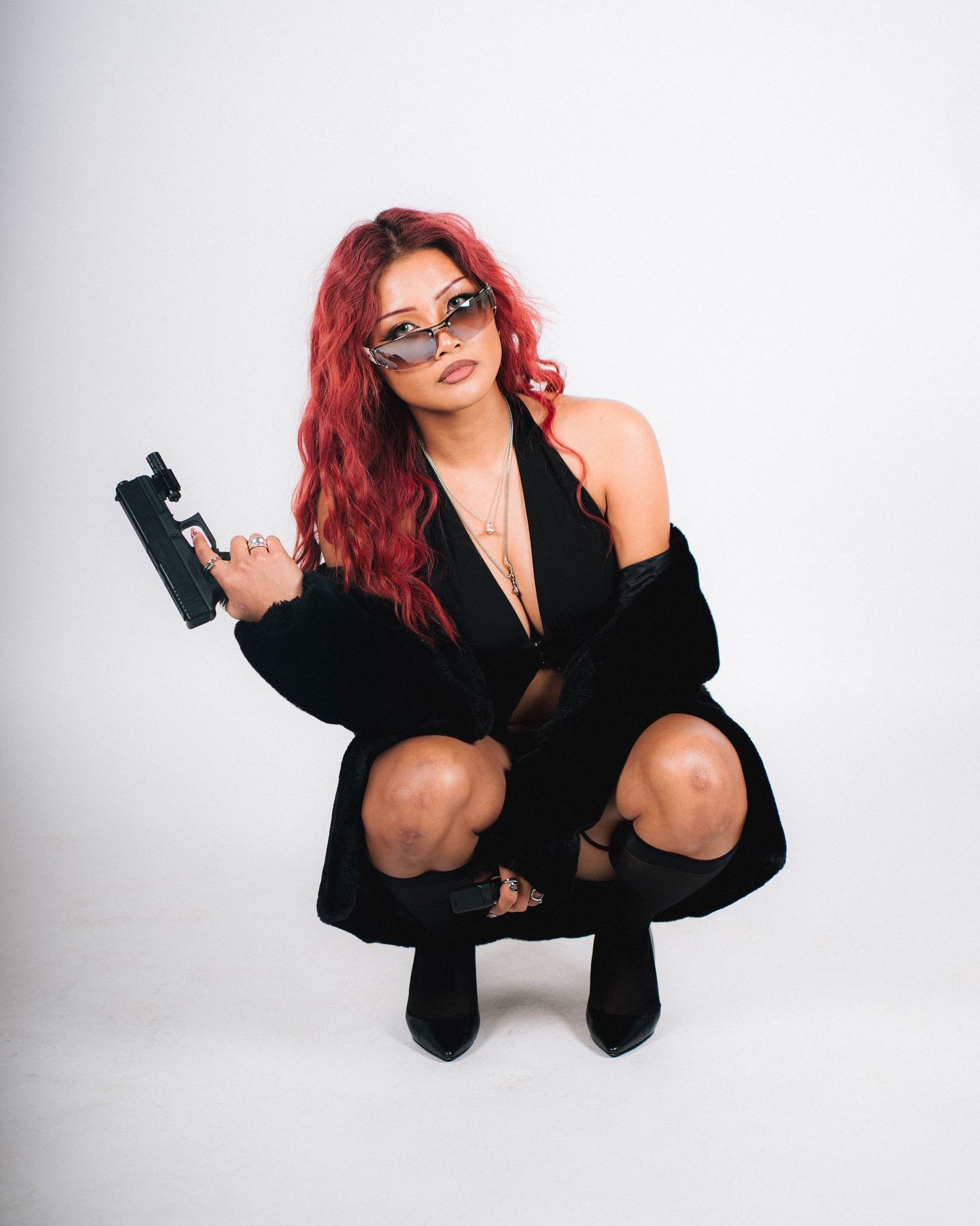 A fashion portrait featuring a person with red hair and glasses squatting in a black outfit against a white background.