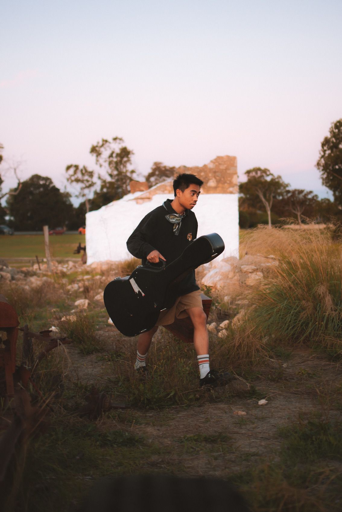 A guitar player in black attire poses outdoors against a rustic backdrop during sunset with tall grass and trees.