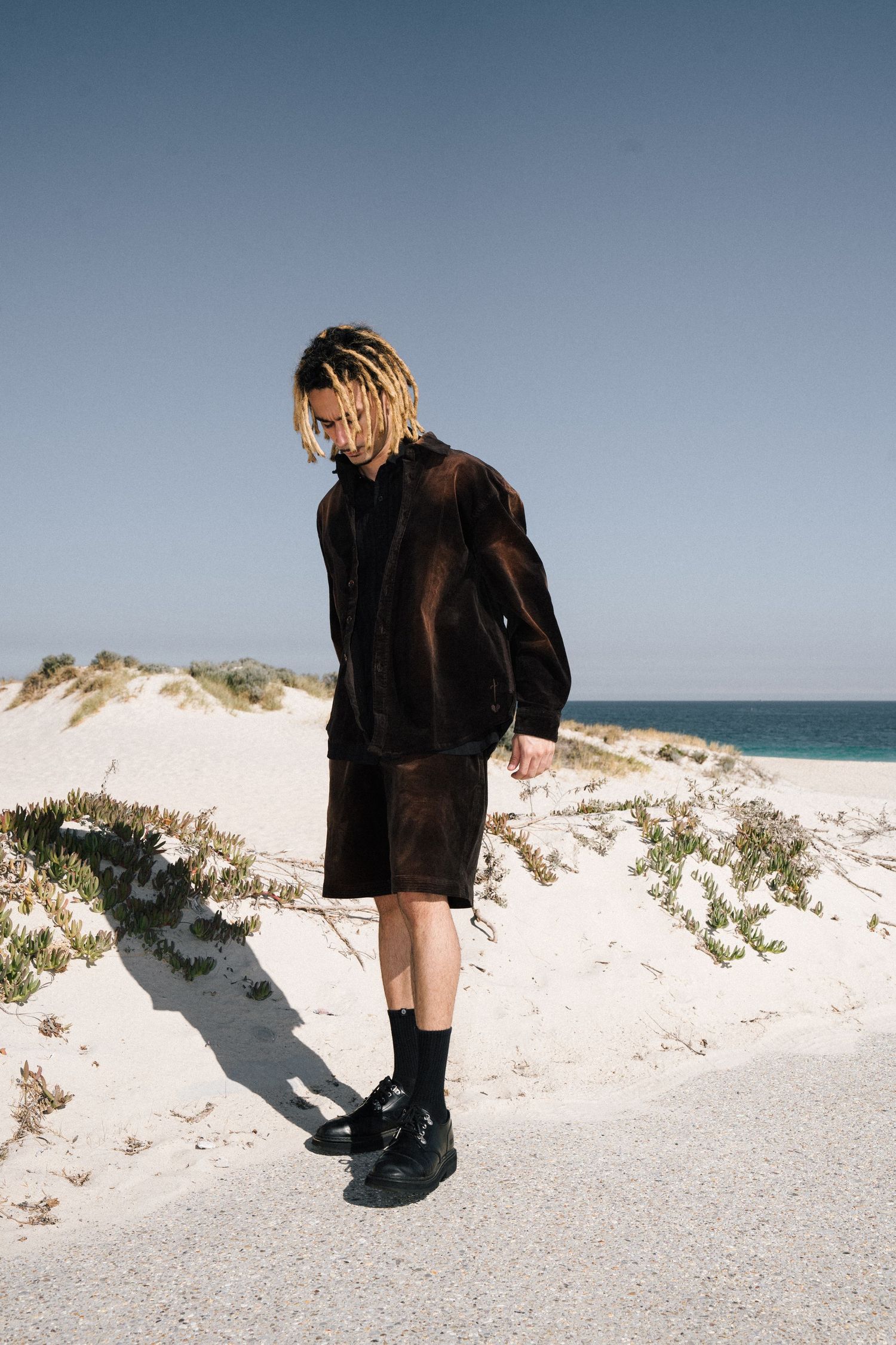 Person in black outfit and boots stands on white sand dunes with ocean and blue sky in background.