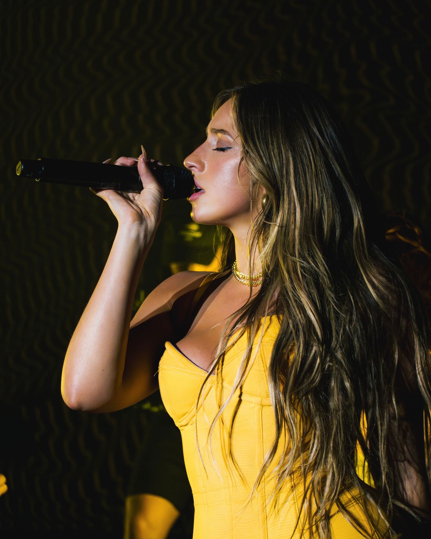 A performer in a yellow dress sings passionately on stage with long flowing hair in dramatic lighting.