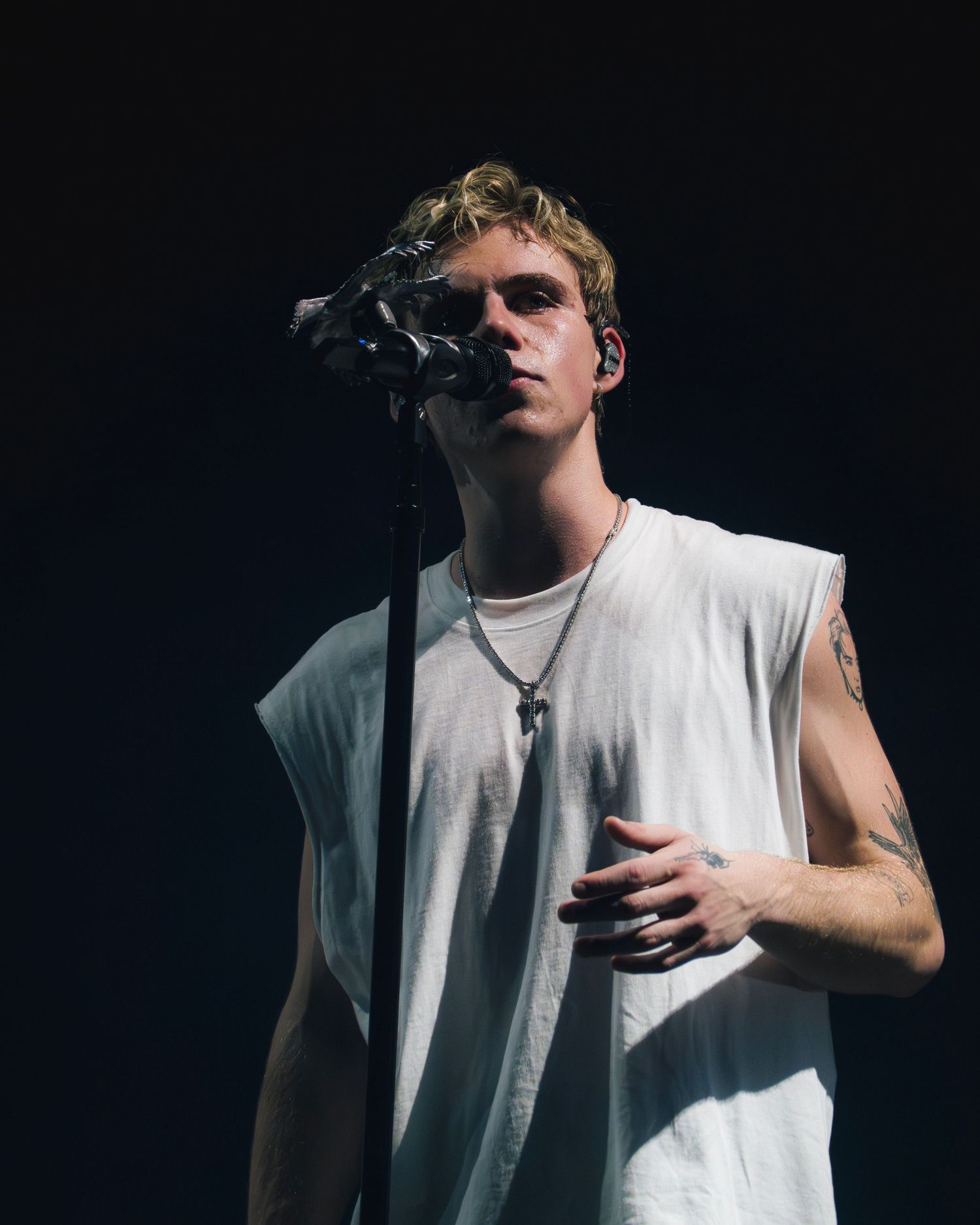 Performer in a white sleeveless shirt singing on stage against a dark background during a dramatic performance.
