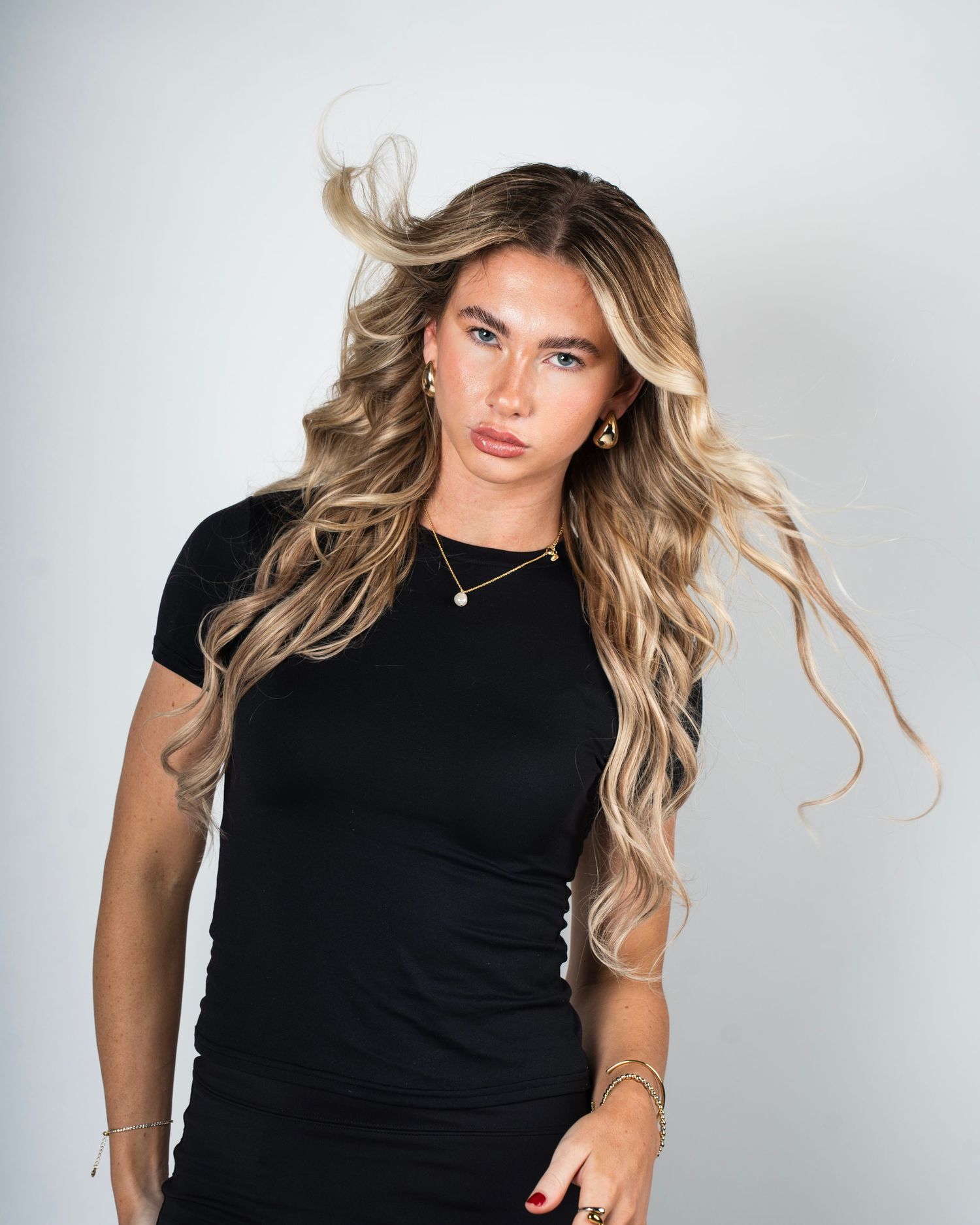 Woman in black t-shirt with long wavy blonde hair posing against white background.