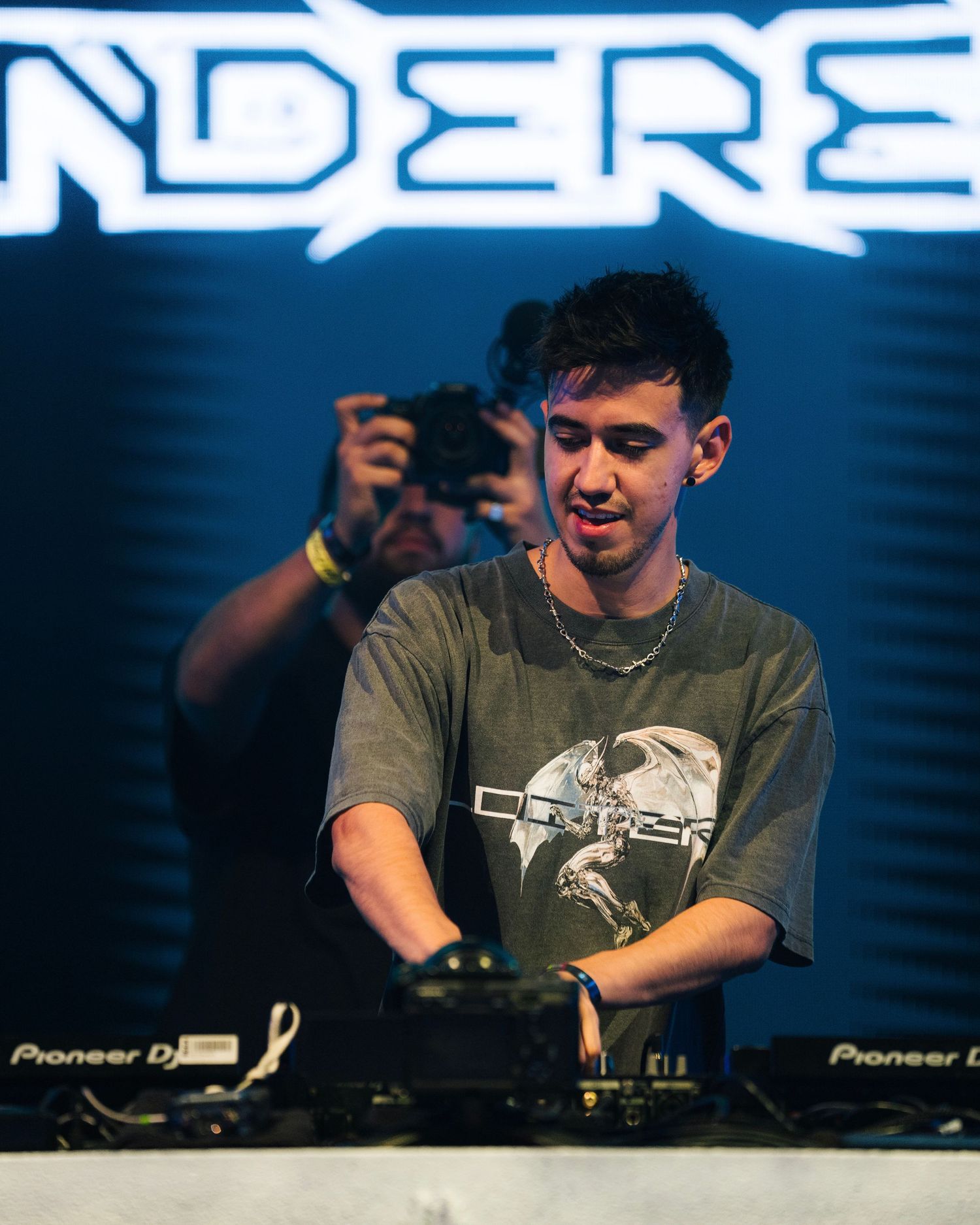 DJ performs at electronic music event with blue LED signage in background.