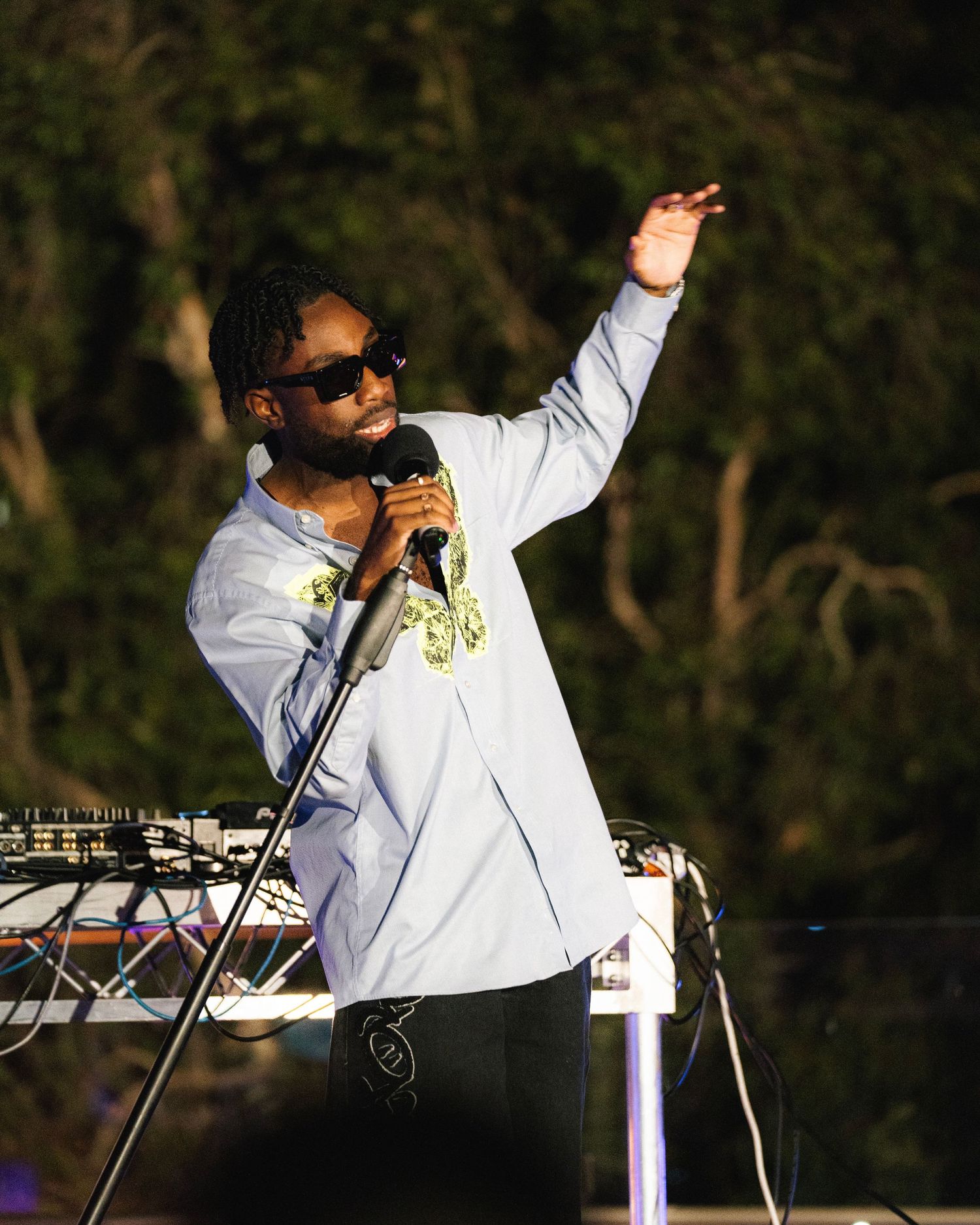 Performer in white sweatshirt and sunglasses singing into microphone on outdoor stage with raised arm gesture.