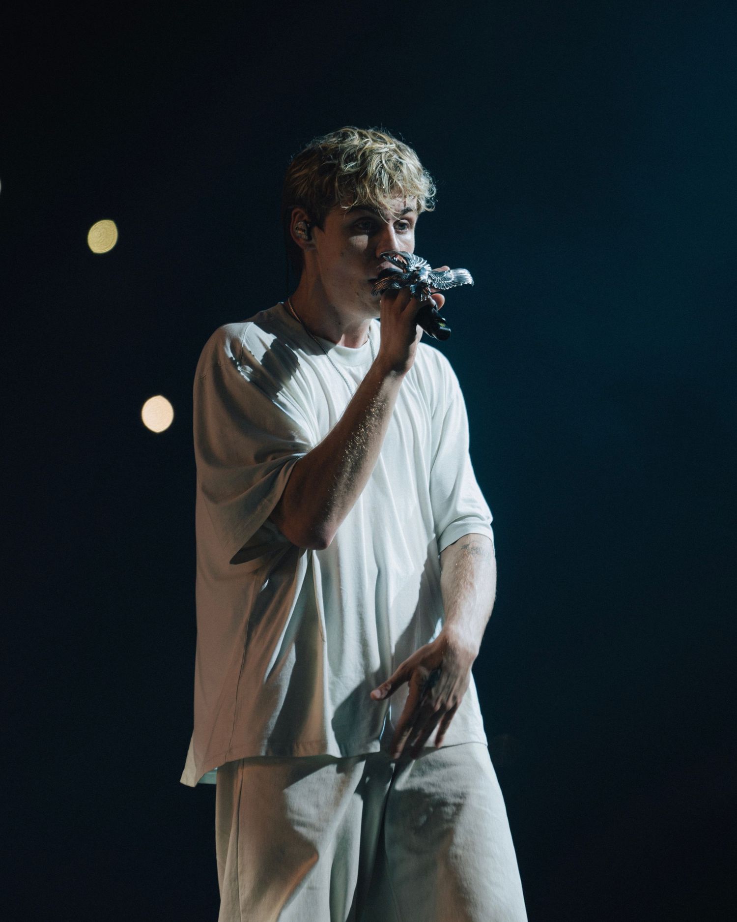Concert performer in white outfit singing into microphone against dark background with stage lighting.
