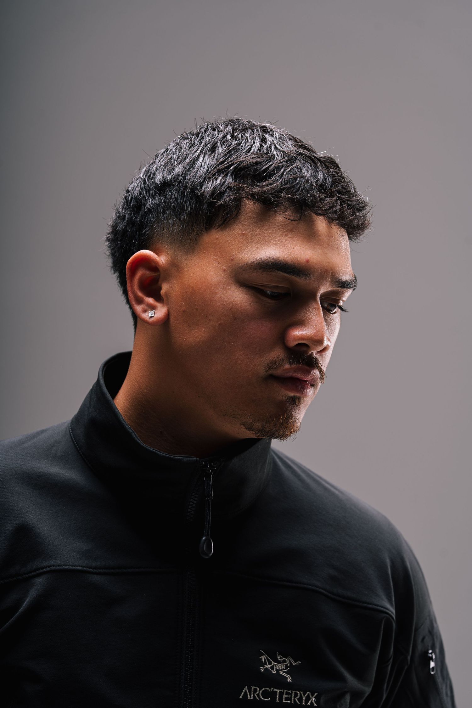 Profile view of a person wearing a black Arc'teryx jacket and wireless earbud in a contemplative pose against a gray background.