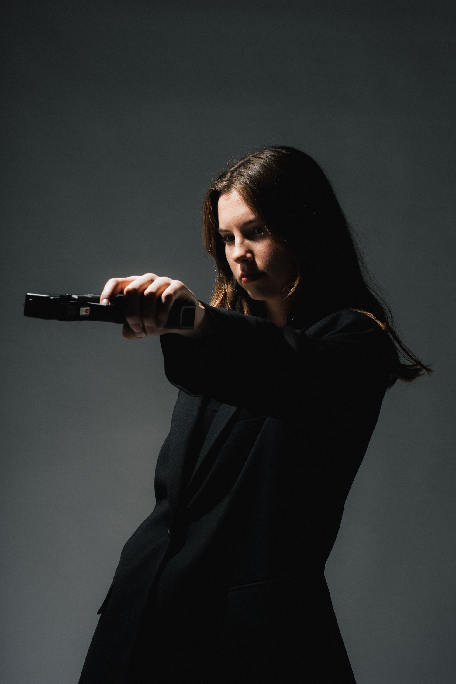 Dramatic silhouette of someone in dark clothing holding a gun against moody gray backdrop.