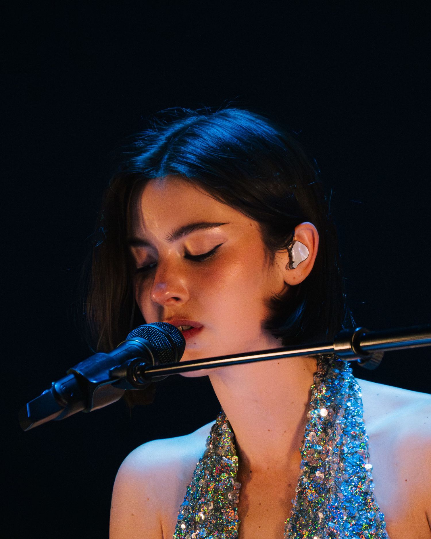Singer performing on stage wearing a sparkly dress while holding a microphone in a darkly lit concert venue.