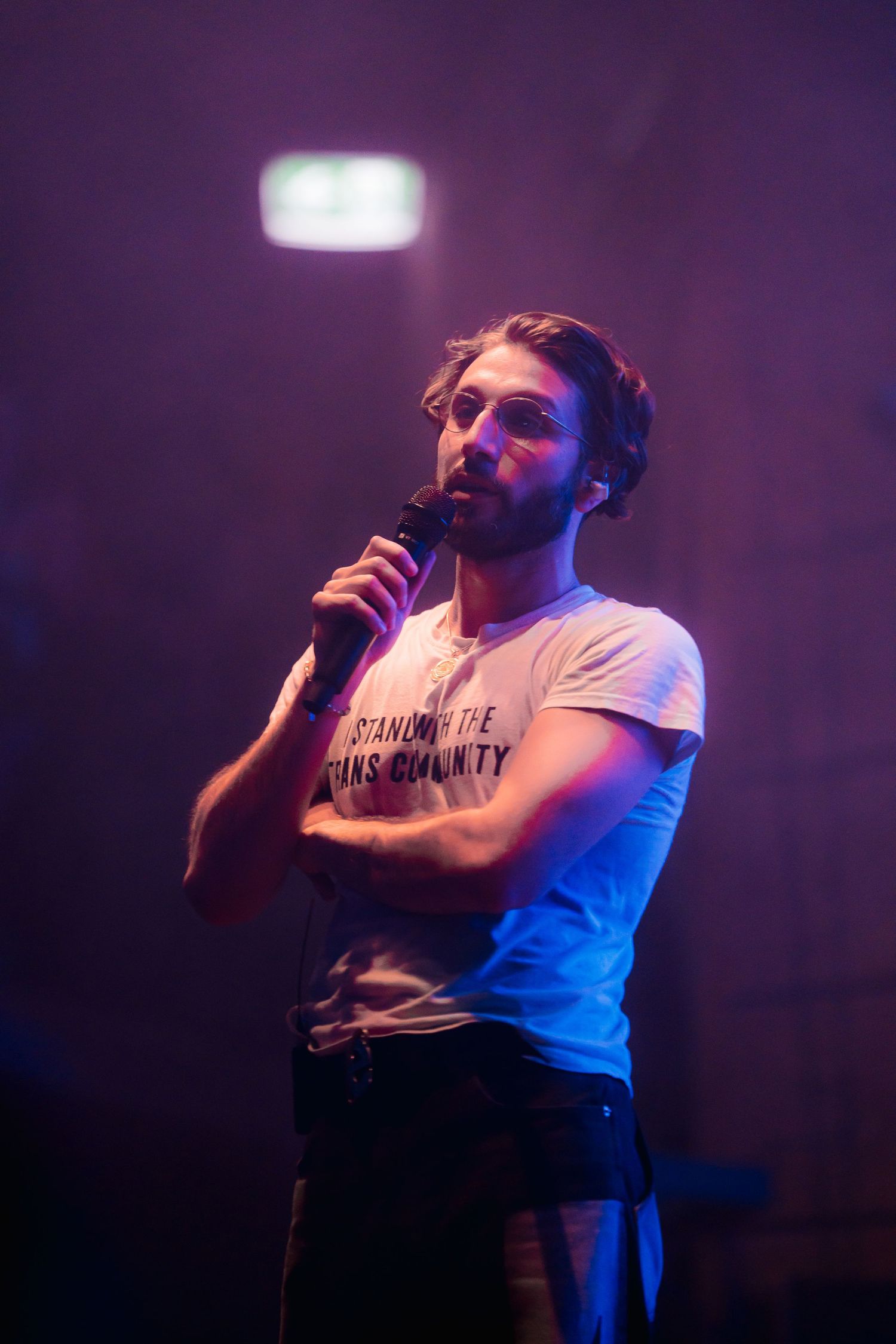 A performer in a white t-shirt sings into a microphone on stage with purple lighting in the background.